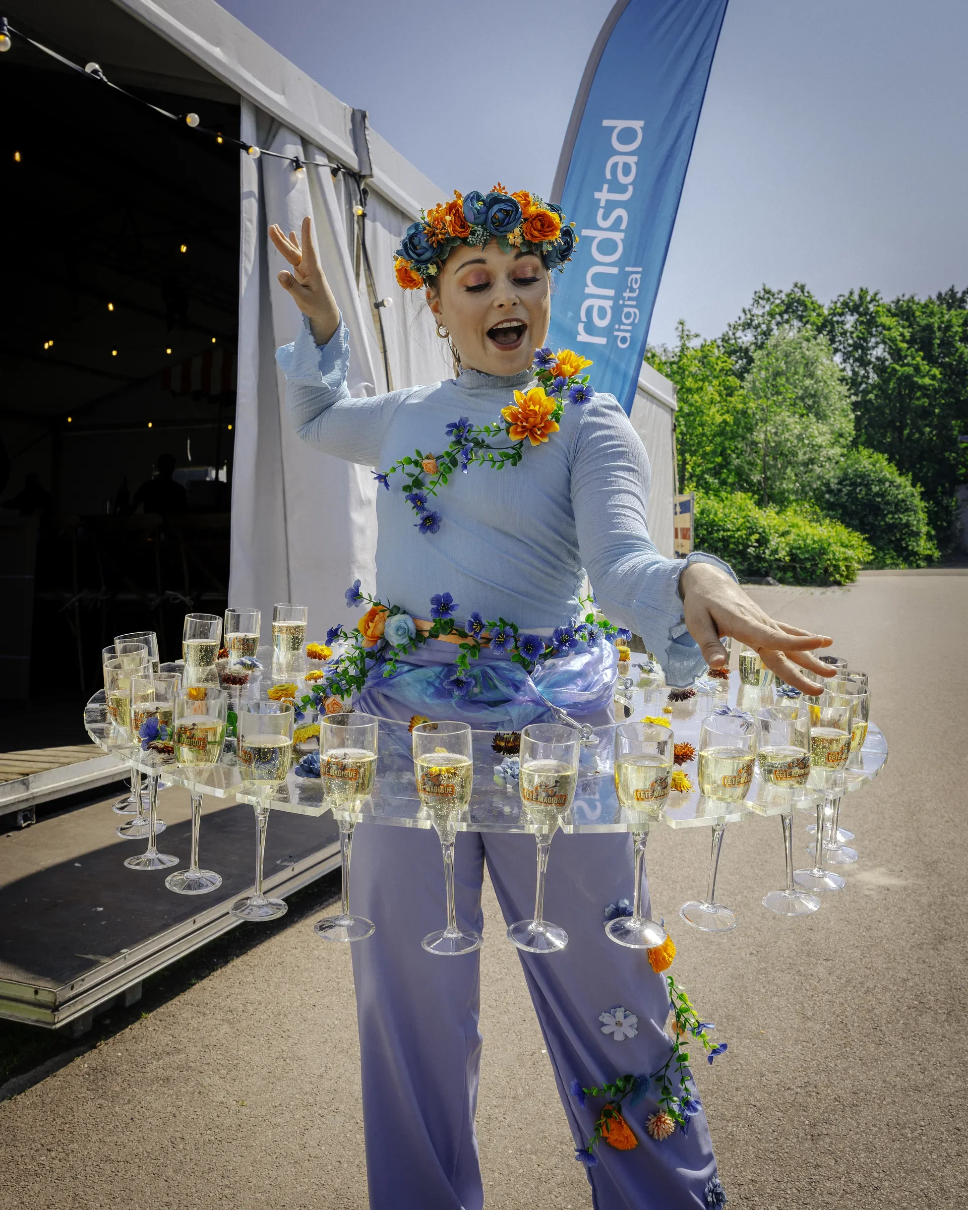 Persoon in een blauwe jurk met een bloemkroon danst bij een tafel met glazen champagne, buiten onder een blauwe vlag met de tekst 'randstad digital'.