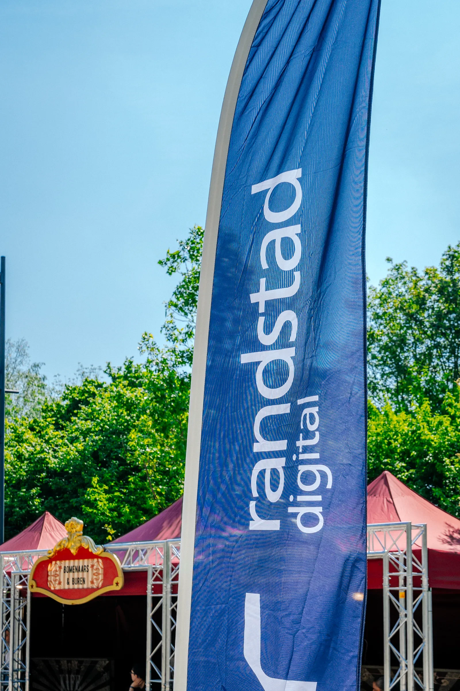 Een blauwe vlag met witte tekst die gedeeltelijk zichtbaar is, in een buitenomgeving met groene bomen en een blauwe lucht, en een attractie met een rood dak en een attractie met een bord die zegt 'Bomenars & Buren'.