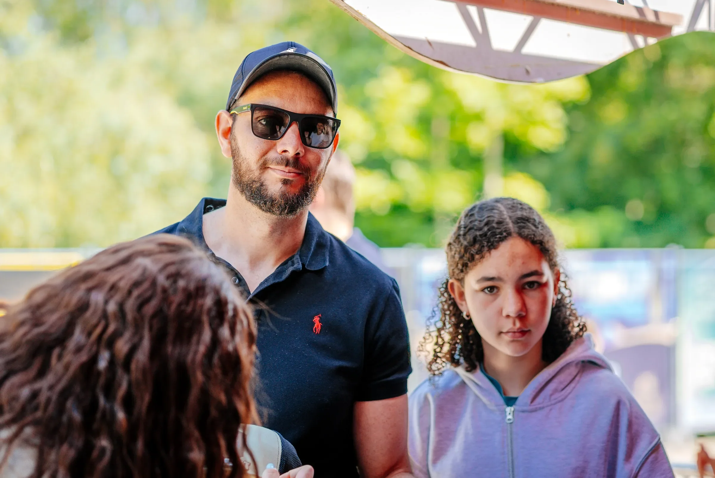 Een man met een pet en zonnebril praat met een groep kinderen buiten op een groene plek.