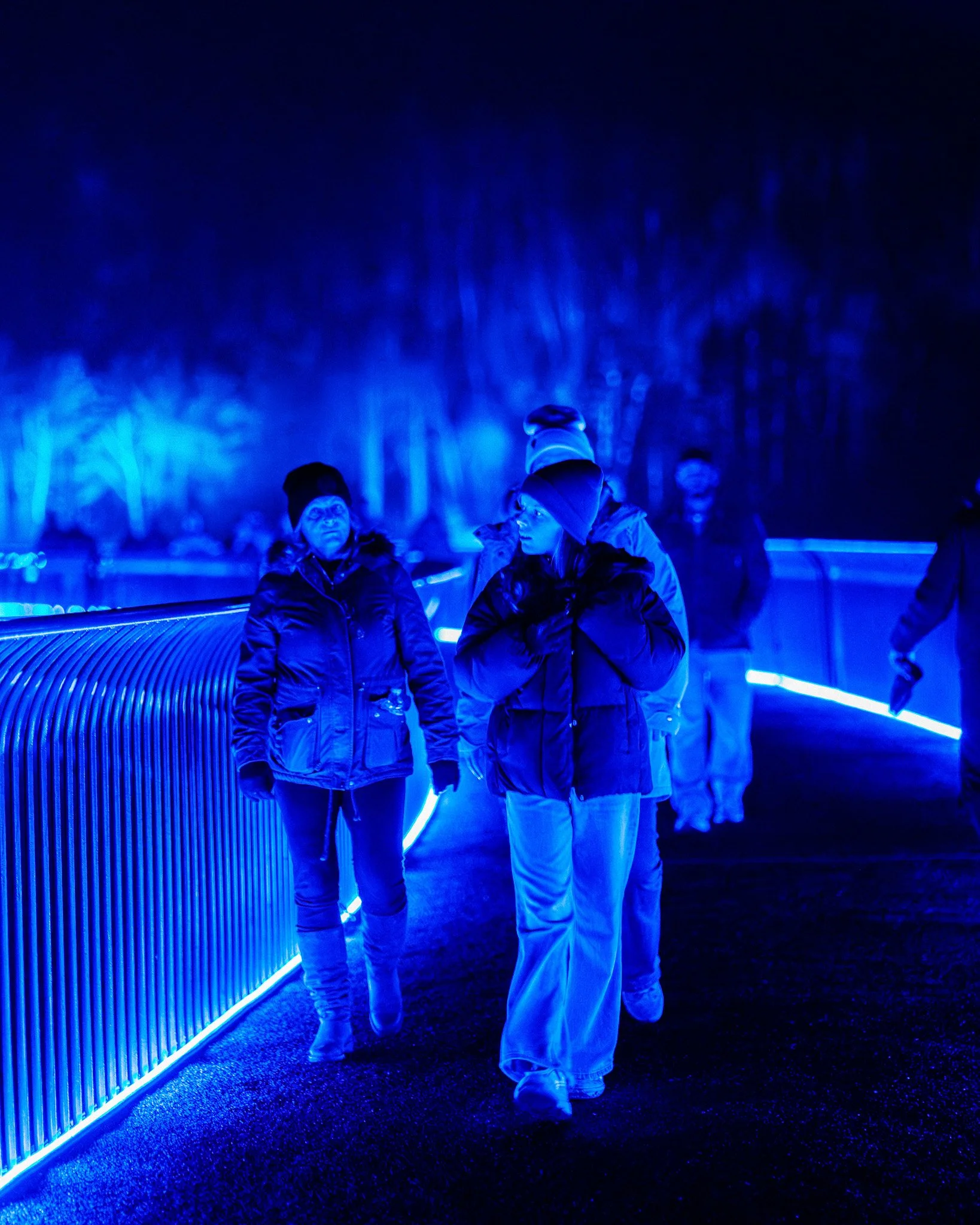 Mensen die lopen in een donker, blauw verlichte omgeving met een metalen rand en lichte lijnen op de vloer.