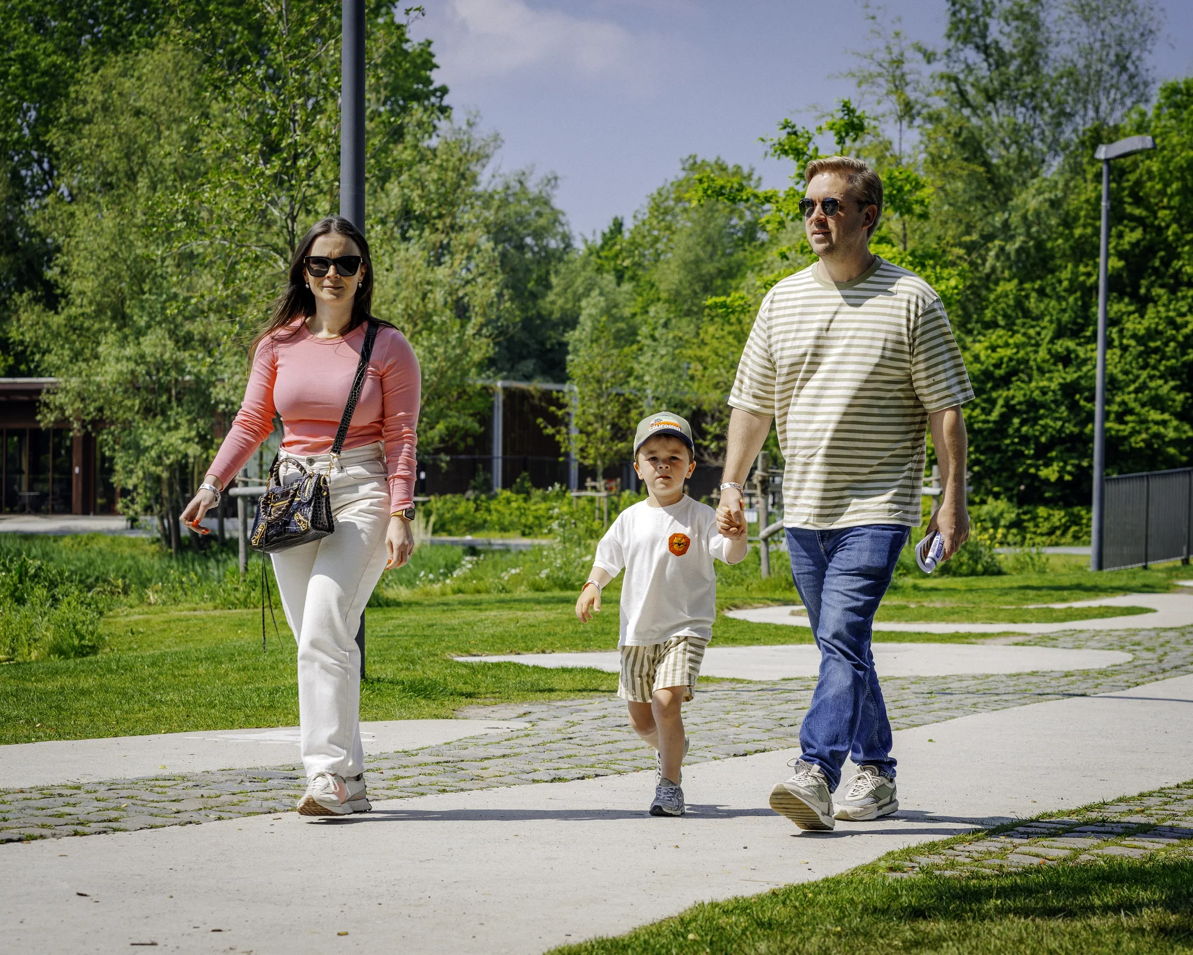 Een familie wandelt door een park op een zonnige dag, bestaande uit een man, een vrouw, een jongen en een baby.