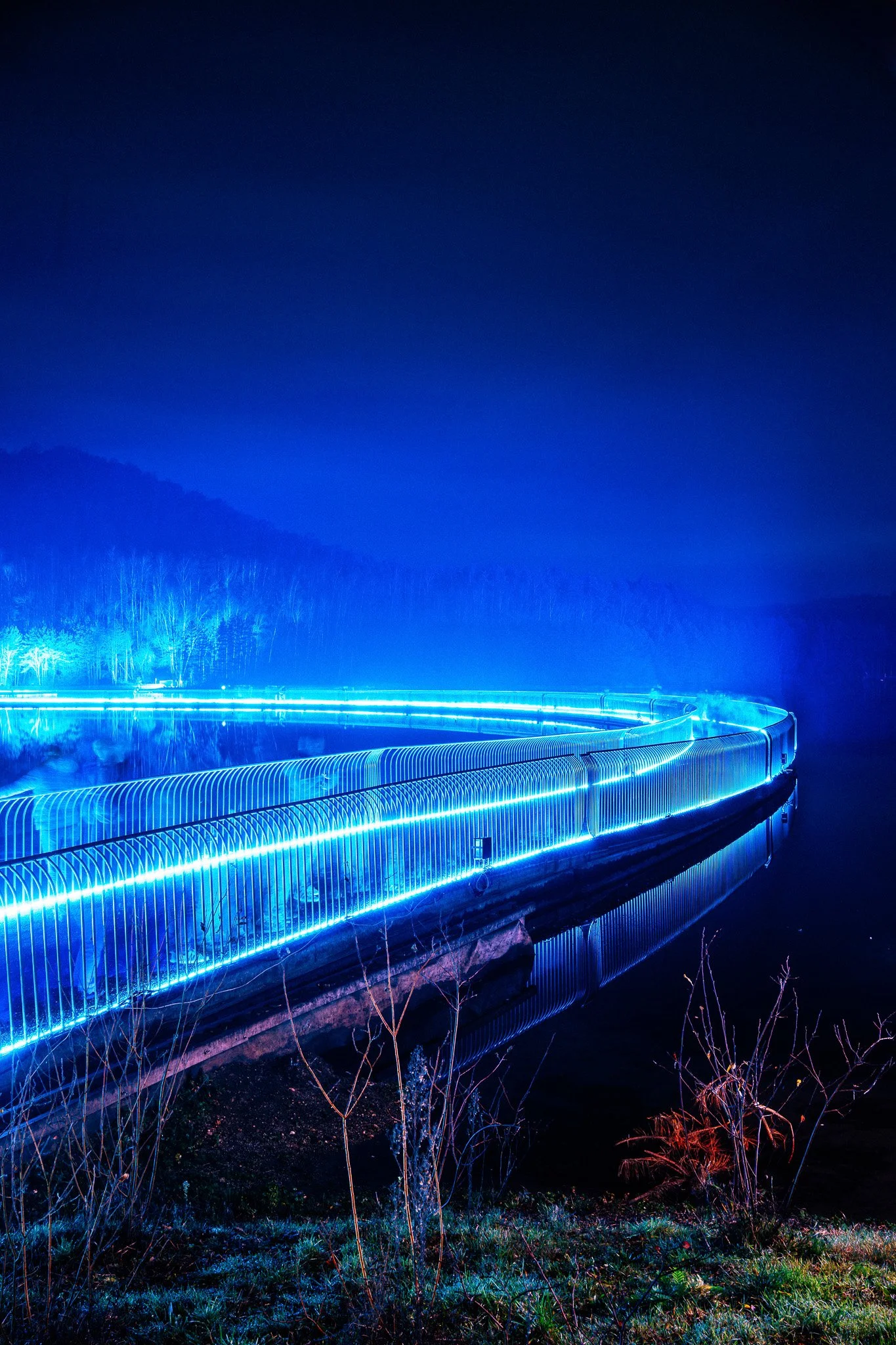 Een verlichte brug met blauwe neonlichten in een landelijke omgeving 's nachts, omgeven door gras en bomen.