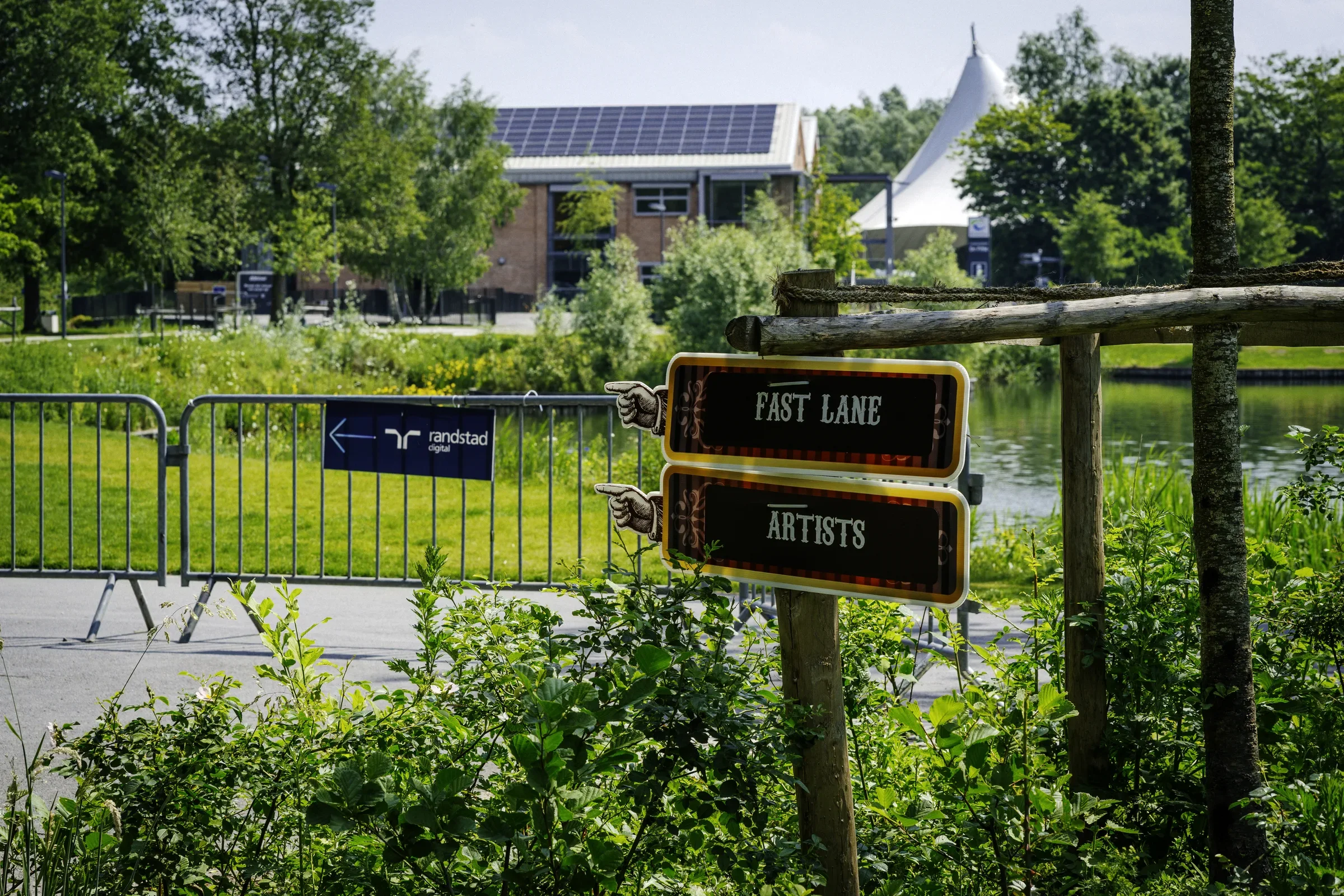 Een bord met de tekst 'Fast Lane' en 'Artists' in een groene omgeving naast een rivier, met een gebouw met zonnepanelen op het dak op de achtergrond.