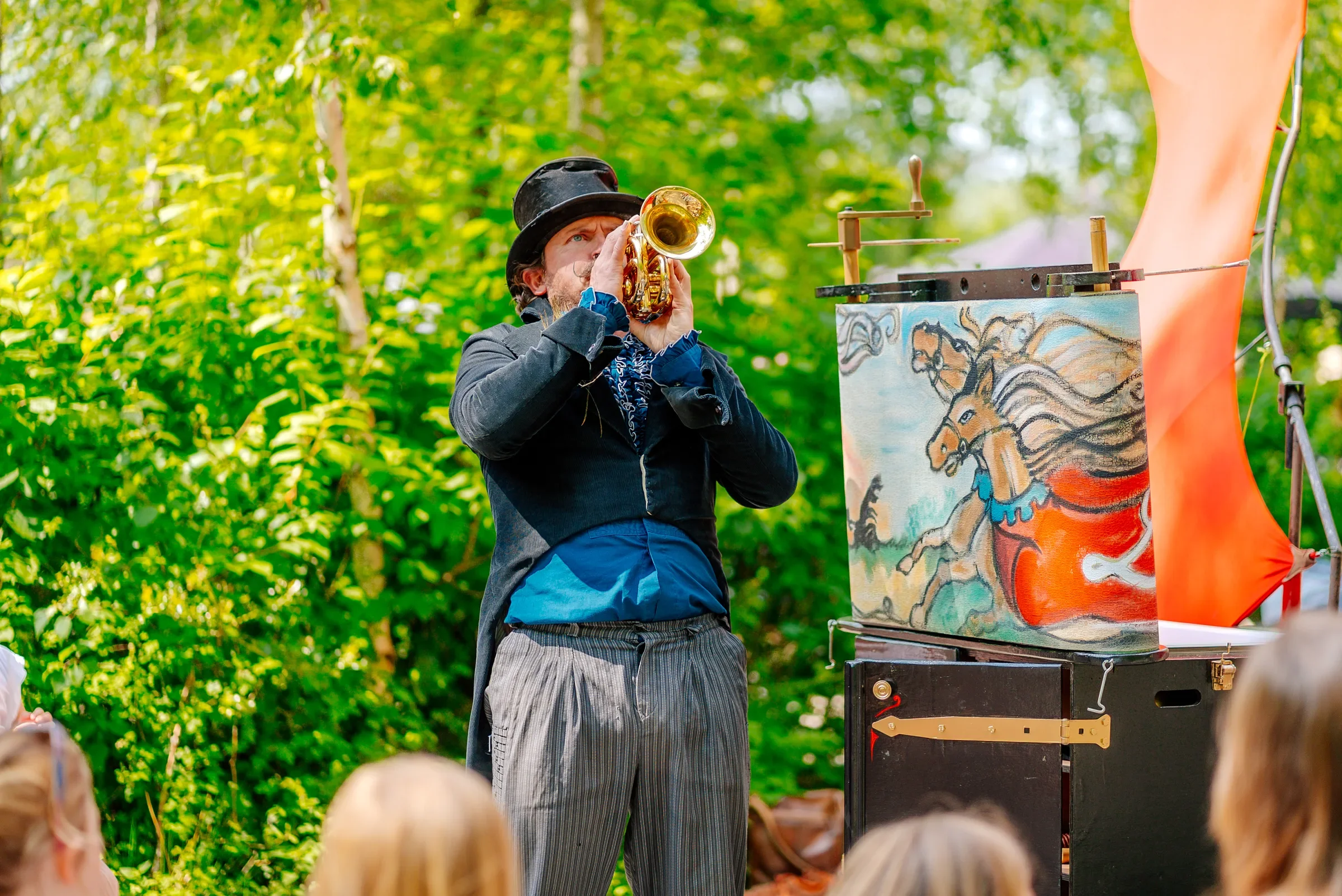 Man in eenhoornkostuum die met een klein trompetje speelt op een buitenpodium tijdens een evenement, omringd door een publiek in een bosrijke omgeving.