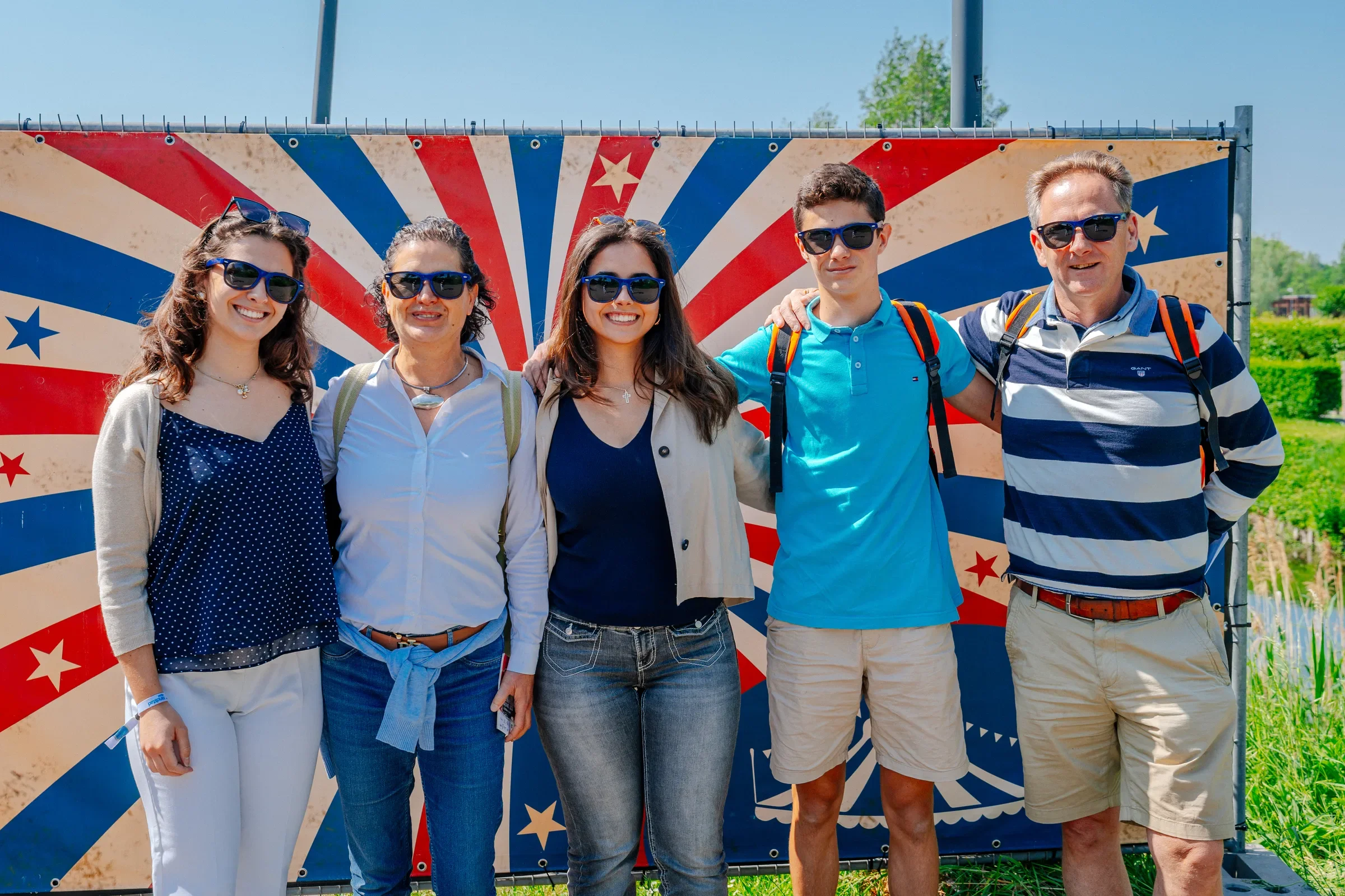 Vijf personen in Zomerkleding met zonnebrillen staan voor een kleurrijke achtergrond met sterren- en streeppatroon, waarschijnlijk tijdens een buitenactiviteit of festival.