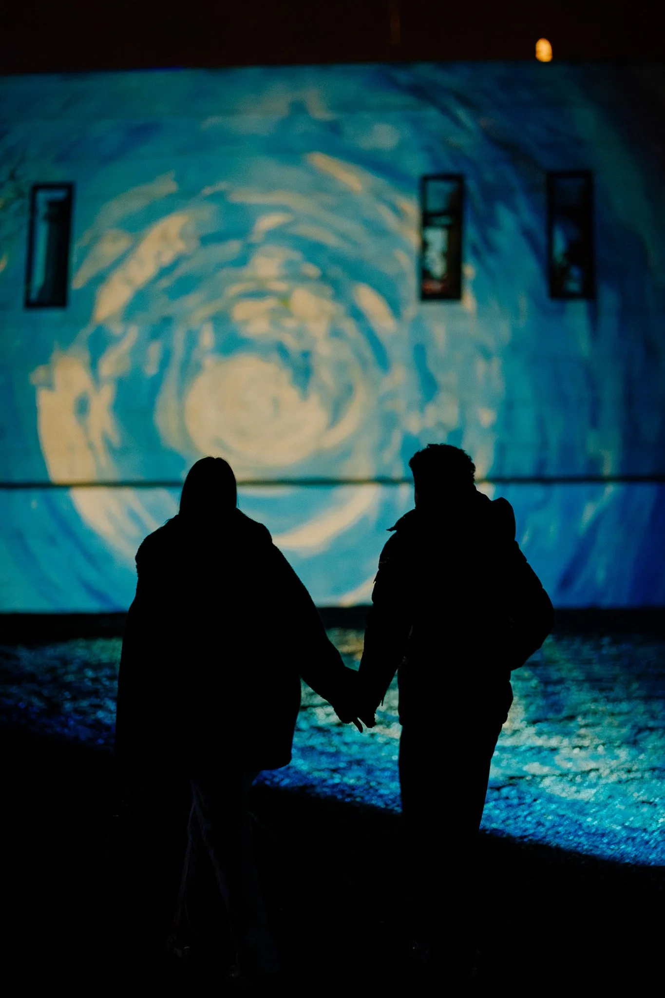 Twee mensen die op de oever van een meer staan en een grote projectiescherm bekijken met blauwe en gele kleuren, mogelijk een kunstinstallatie of filmprojectie, tijdens de nacht.