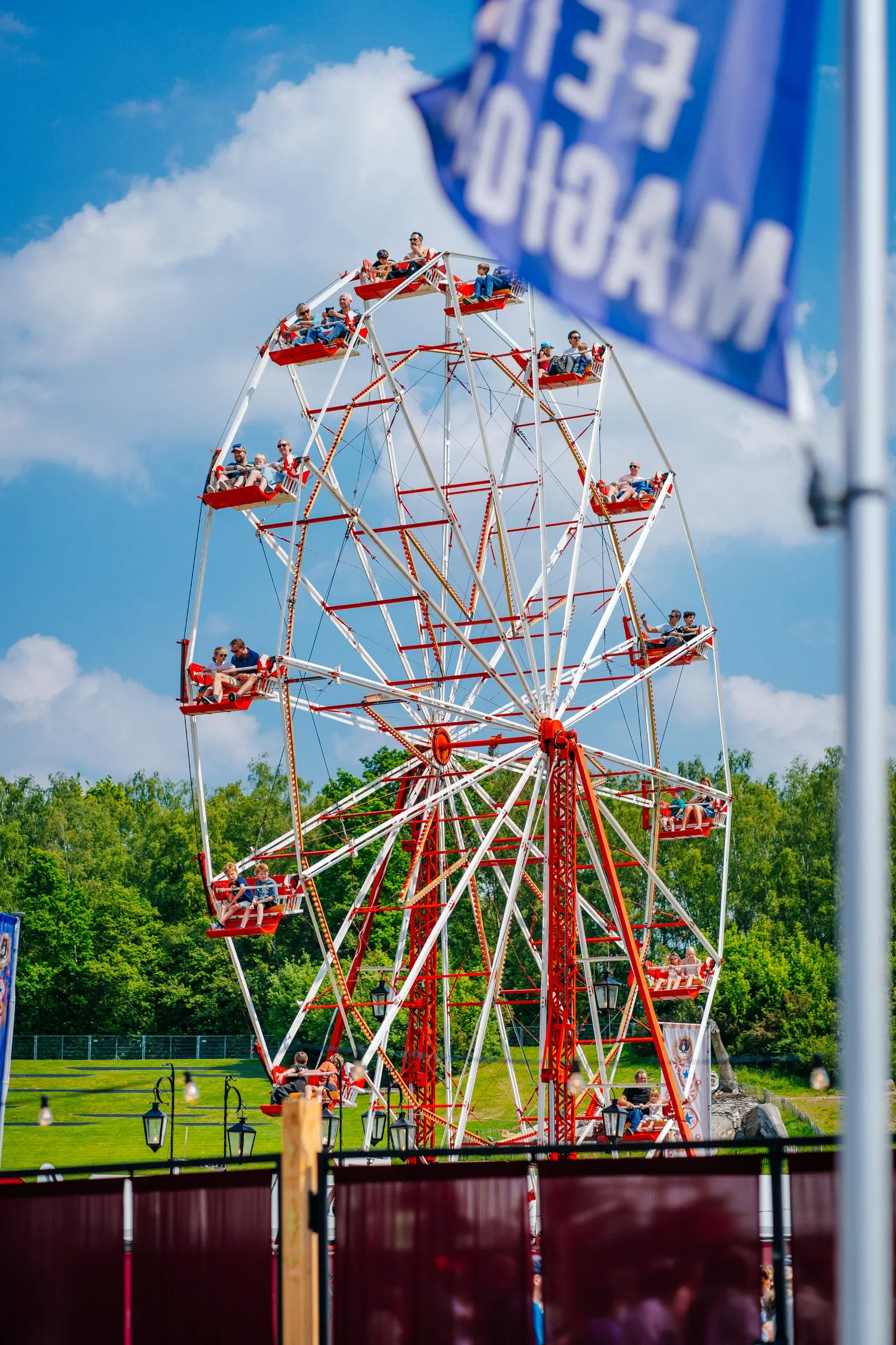 Een grote rode wheel of fortune op een pretpark met mensen die erin zitten, blauwe lucht en groene bomen op de achtergrond.