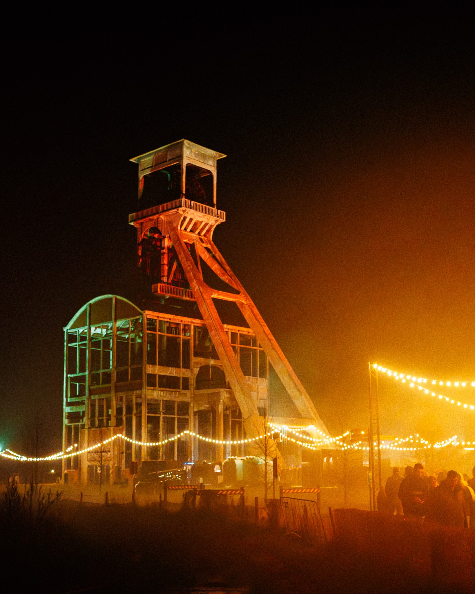 Een attractiepark of kermis tijdens de avond met een grote houten glijbaan en gekleurde verlichting, mensen die eromheen staan en lopen.