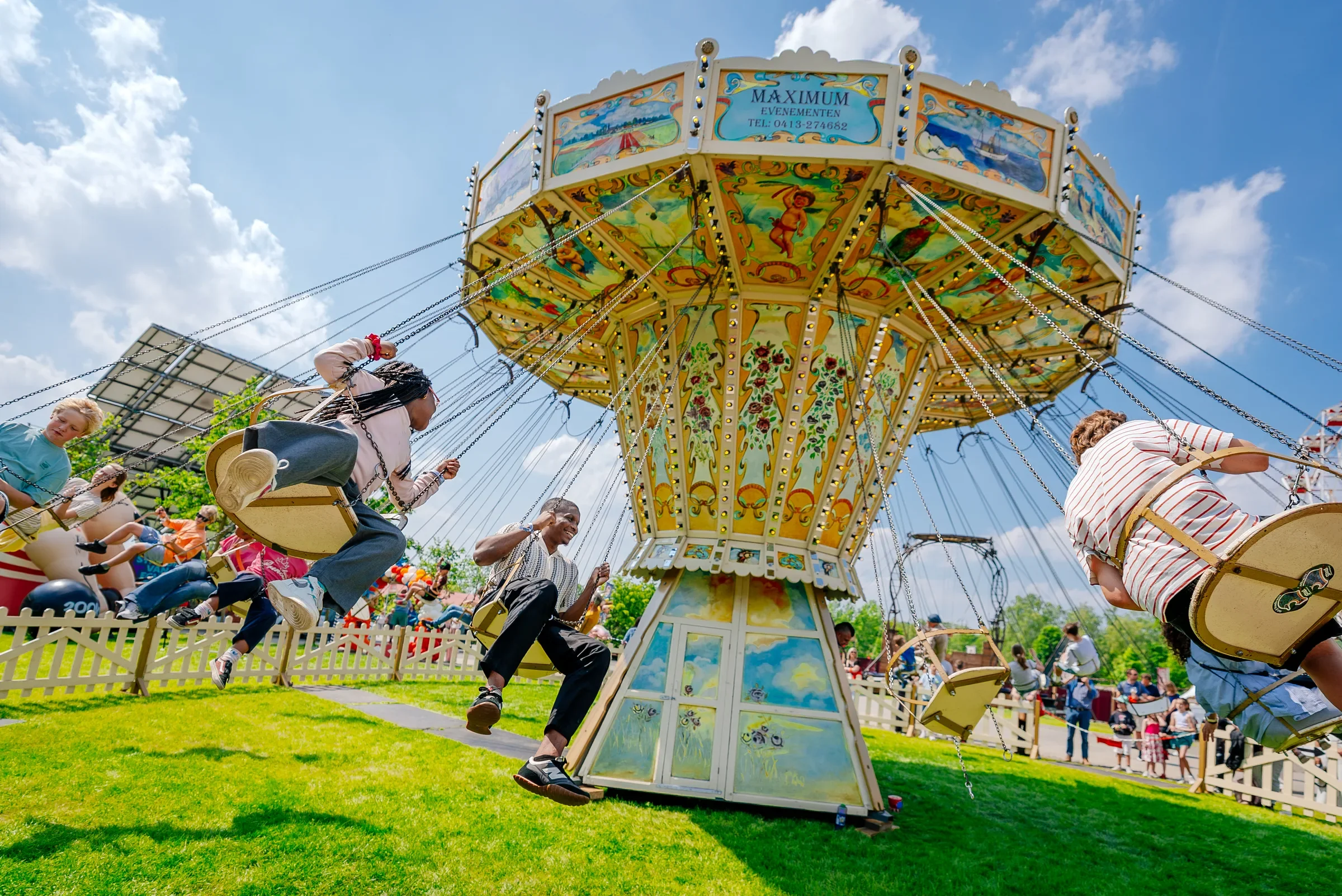 Mensen genieten van een draaiend vrolijk wiel op een kermis, met een blauwe lucht en witte wolken op de achtergrond.