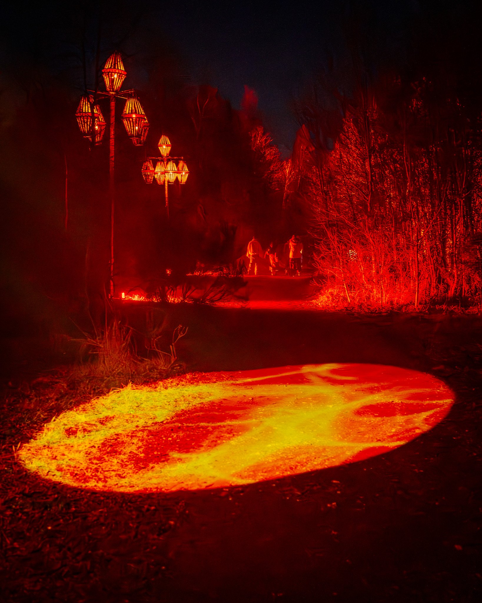 Lava met rode en gele kleuren stroomt door de donkere bosomgeving met bomen. Er zijn enkele personen zichtbaar in de achtergrond.