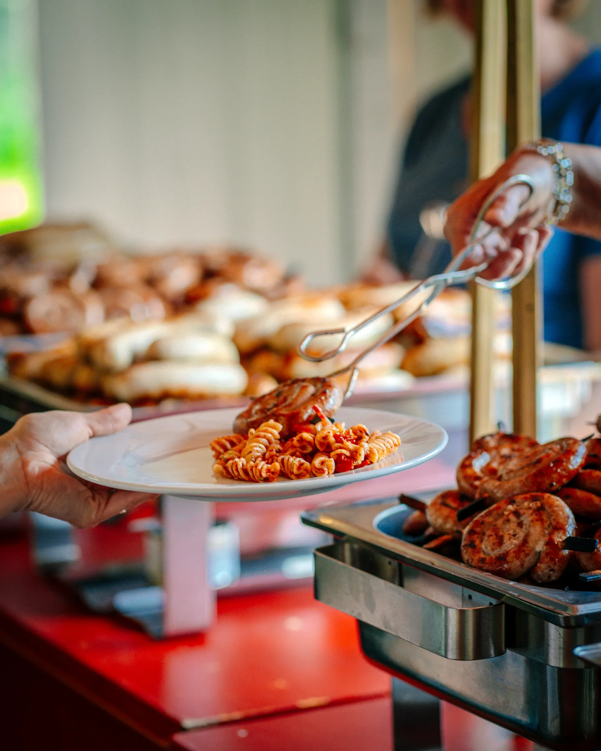 Persoon die pasta op een bord serveert bij een buffet met pizza en gebakken gerechten.