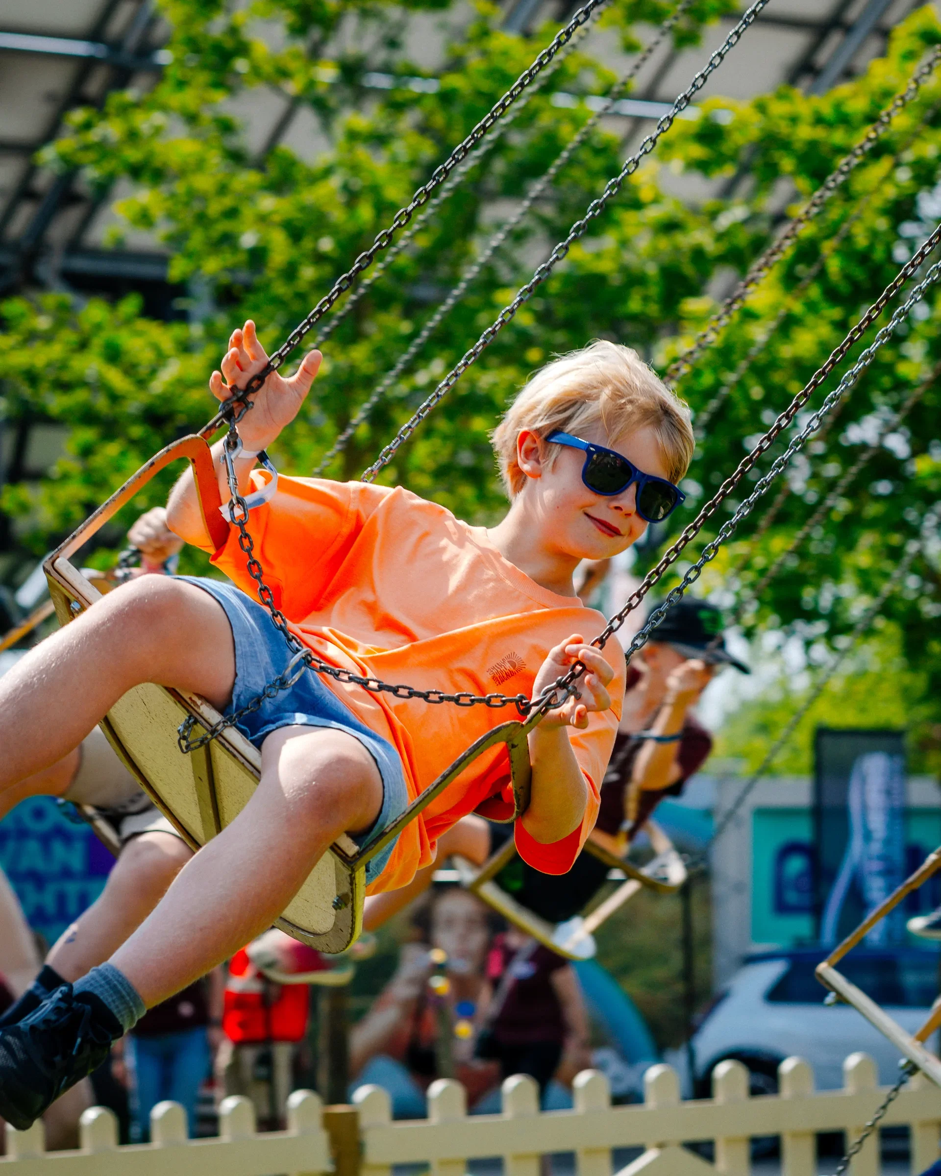 Een kind met blond haar, blauwe zonnebril en een oranje T-shirt zit in een schommelstoel op een kinderspeelplaats, omgeven door andere kinderen en volwassenen.