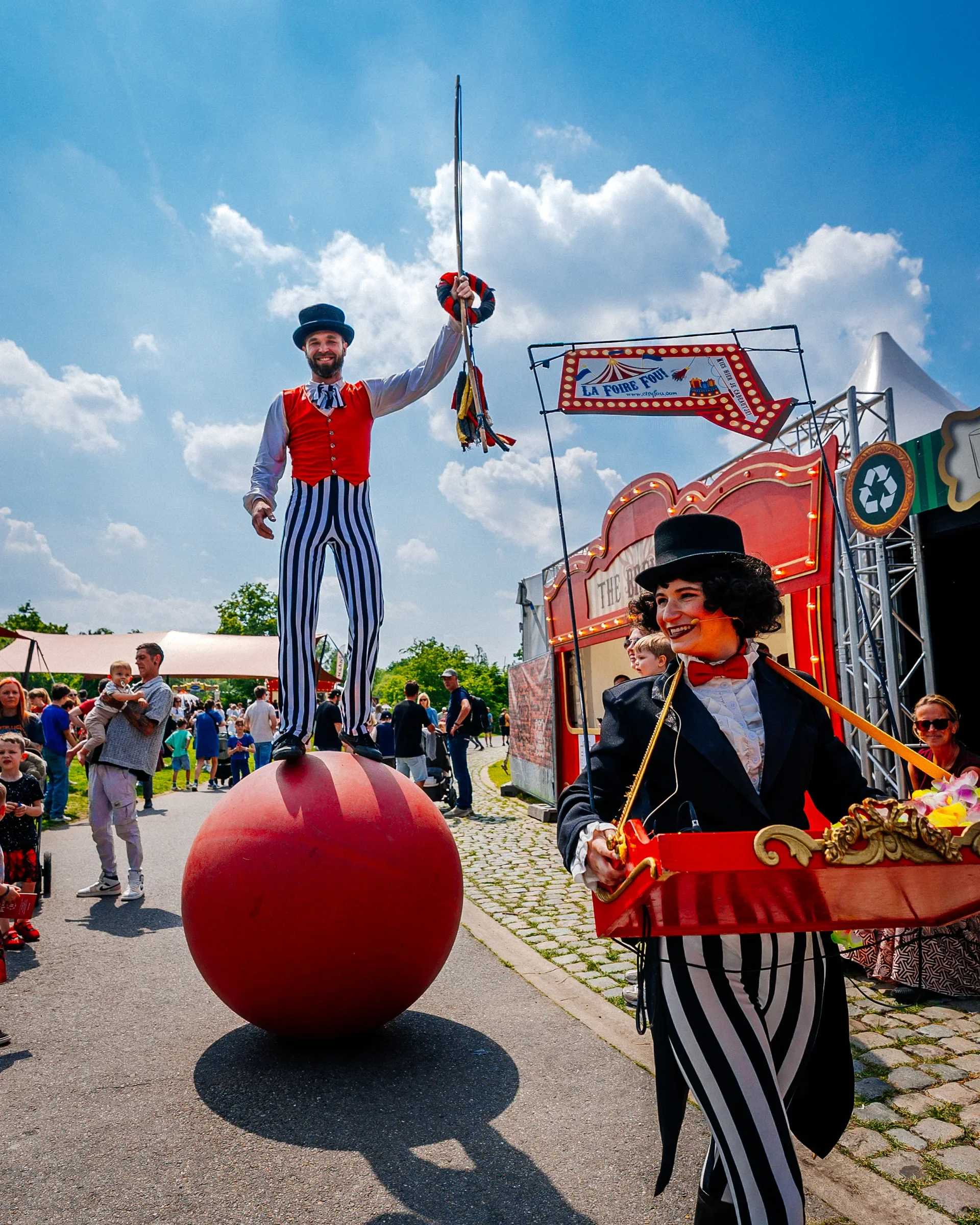 Een man in een circusoutfit met blauwe en witte gestreepte broek en een rode vestje houdt een vlag omhoog tijdens een volksfeest, rondom staan veel mensen en daarachter is een circuswagen met een banner die zegt "La Foire Fou". Er is een grote rode b