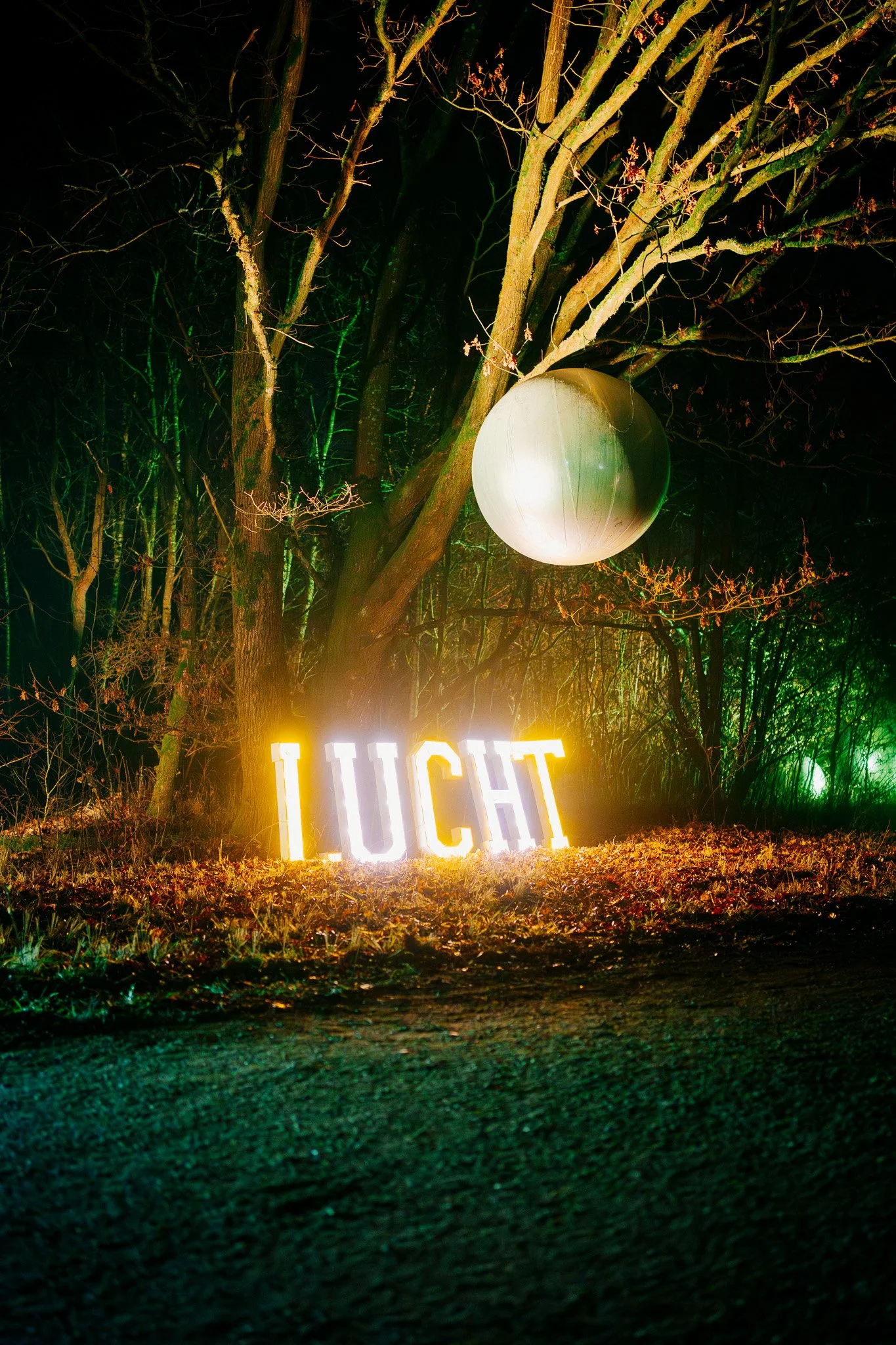 Verlichte letters vormen het woord 'LUCHT' op de grond, met een groot, rond wit lantaarn en een boom met takken en een groene verlichtingsachtergrond in een donker bosgebied.