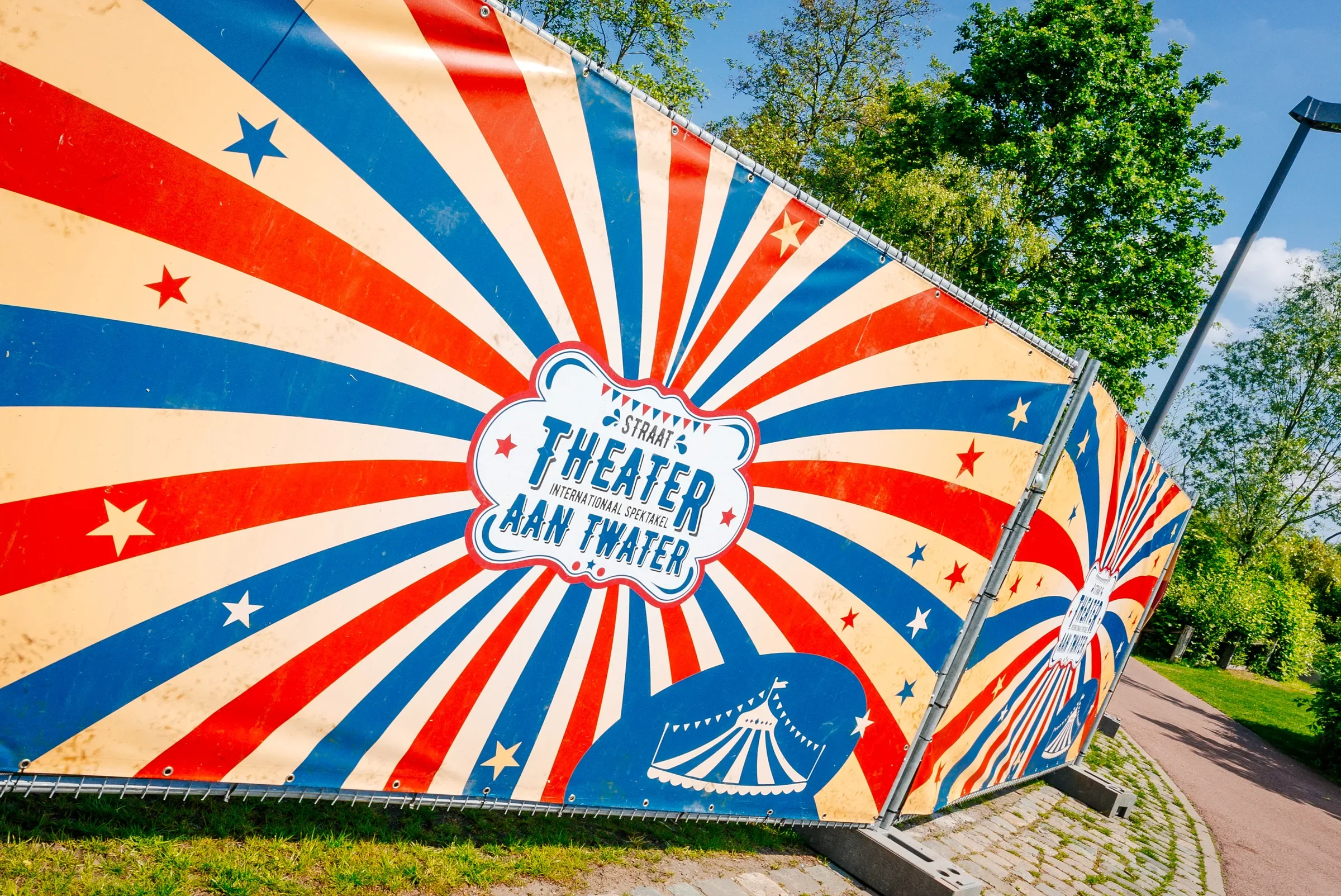 Kermisattractie met rood, wit en blauw design met sterren, met tekst 'Straat Theater' en 'AAN TWITTER' op een tent of scherm, omgeven door groene bomen, onder een blauwe hemel.