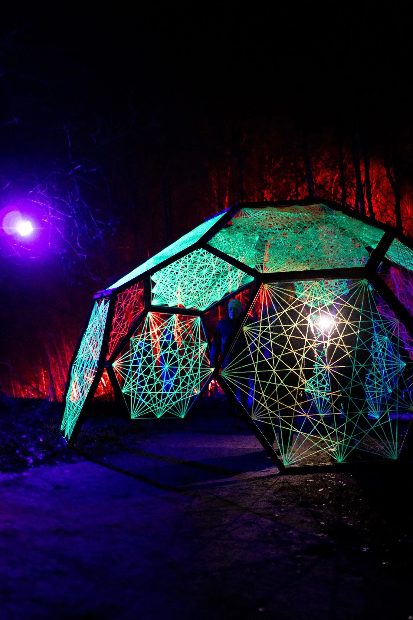Een buitensculptuur van glanzende, geometrisch gevormde structuur met kleurrijke neonlichten in een nachtelijke omgeving, omringd door donkere bomen.