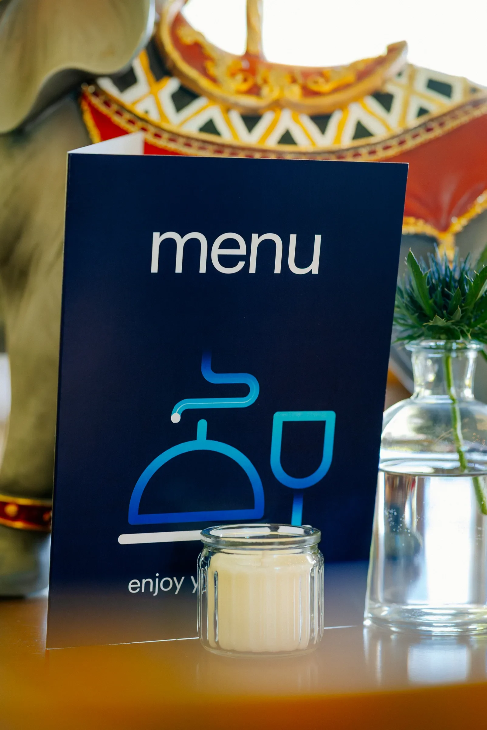 Een menu-kaartje voor een restaurant met de tekst 'menu' en een afbeelding van een kopje koffie en een bord. Er is ook een klein glazen potje met een witte saus of pudding, en een vaas met een groen plantje op de achtergrond.