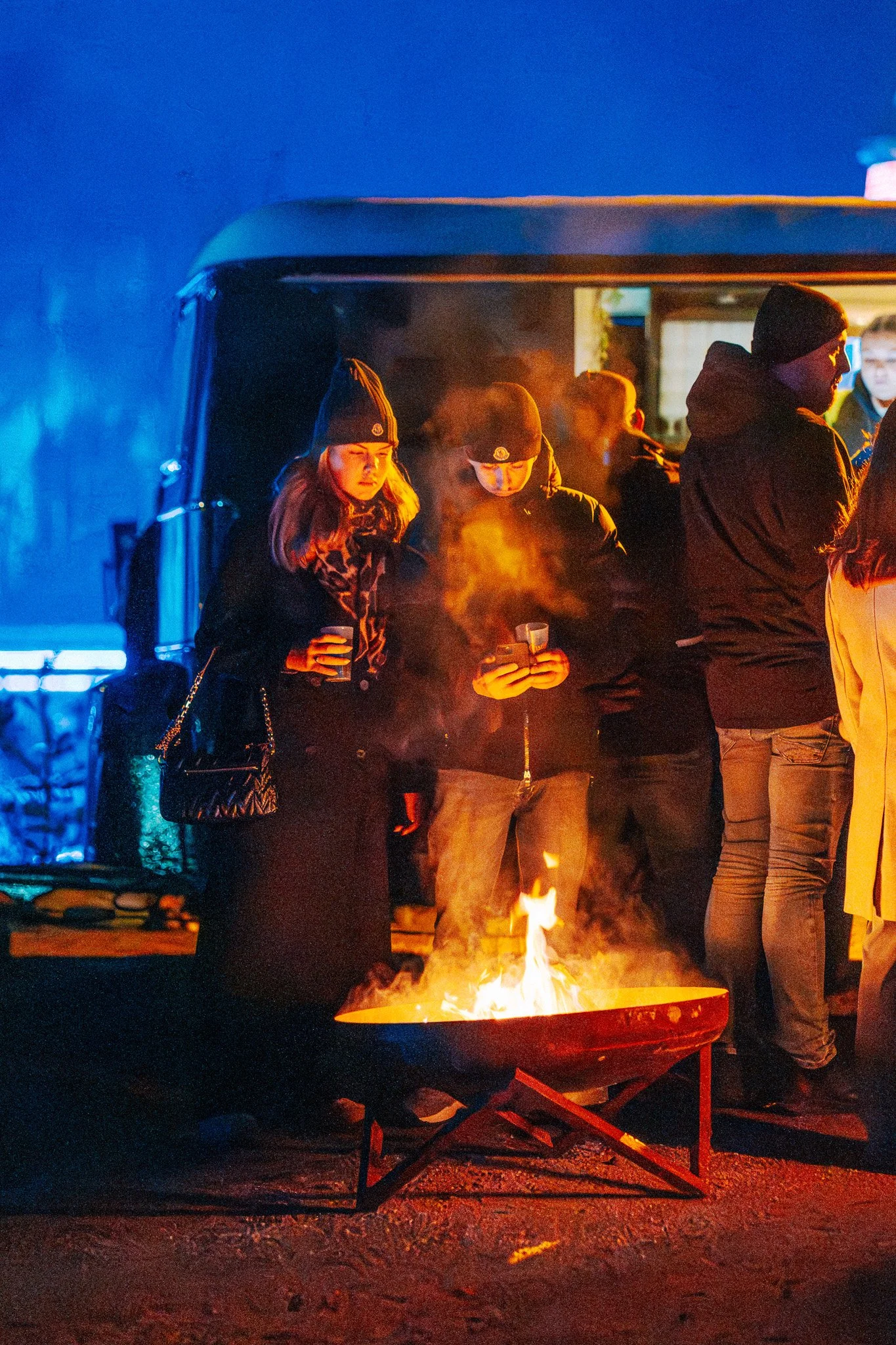 Een groepsfoto van mensen die zich warmen bij een kampvuur 's avonds tijdens een koud weer. Ze dragen winterkleding zoals mutsen en jassen.