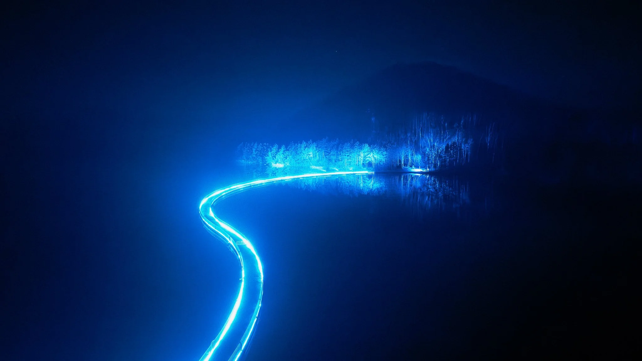 Een nachtlandschap met een smalle, gloeiende, blauwe lichtstrepen die door een waterachtig landschap kronkelen, omgeven door dichtbeboste gebieden en een donkere, mistige atmosfeer.