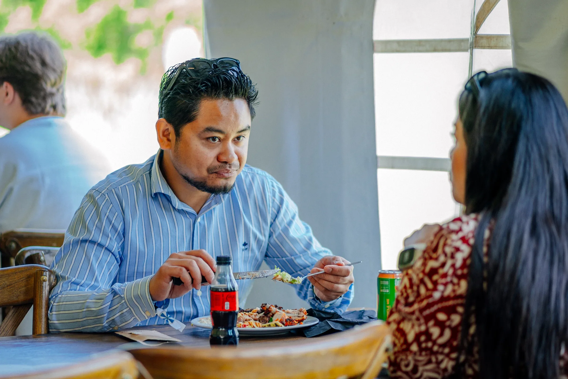 Man met zwart haar en een blauwe en witte gestreepte overhemd eet pizza aan een tafel in een restaurant, met een fles cola en een blikje te zien op de tafel.