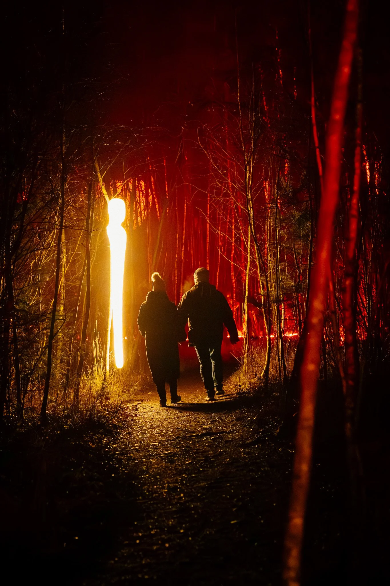 Twee mensen lopen door een donker bos bij nacht, verlicht door een fel, geel-oranje licht en een rood, vurige gloed, met vuurspuwende creatie op de achtergrond.