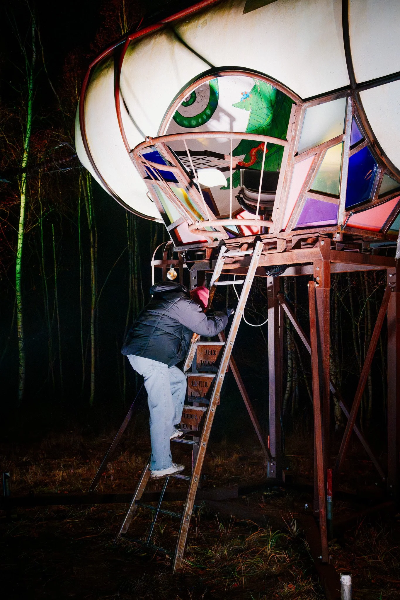 Een persoon die op een ladder staat en naar een grote, kleurrijke, versierde constructie kijkt of deze aanpast. De constructie lijkt op een artistieke structuur met gebruik van metaal en licht, en met een achtergrond van bomen en donkerte.