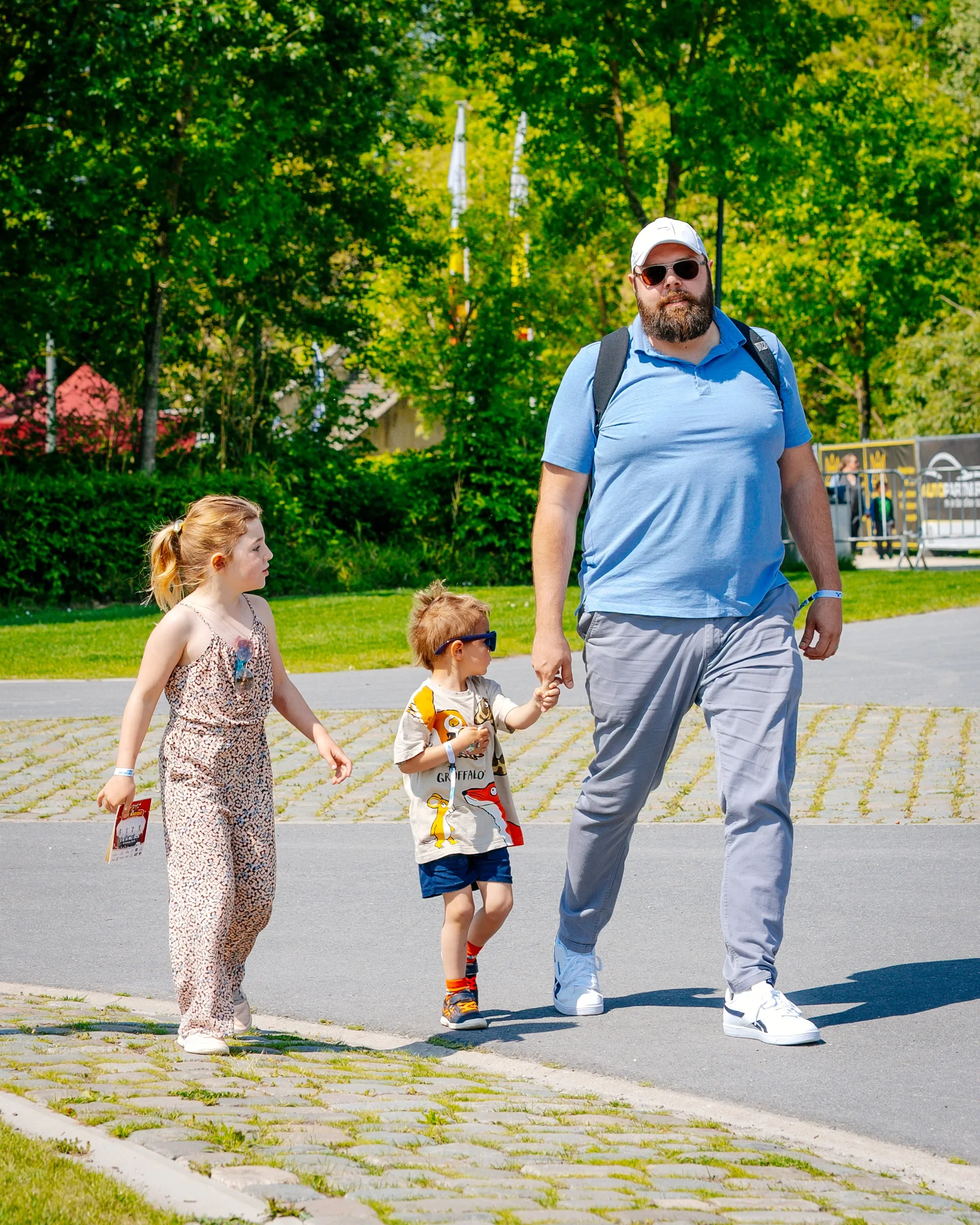 Een man met een grote baard en donkere zonnebril wandelt met drie kinderen op een zonnige dag in een park, omgeven door groen en bomen.