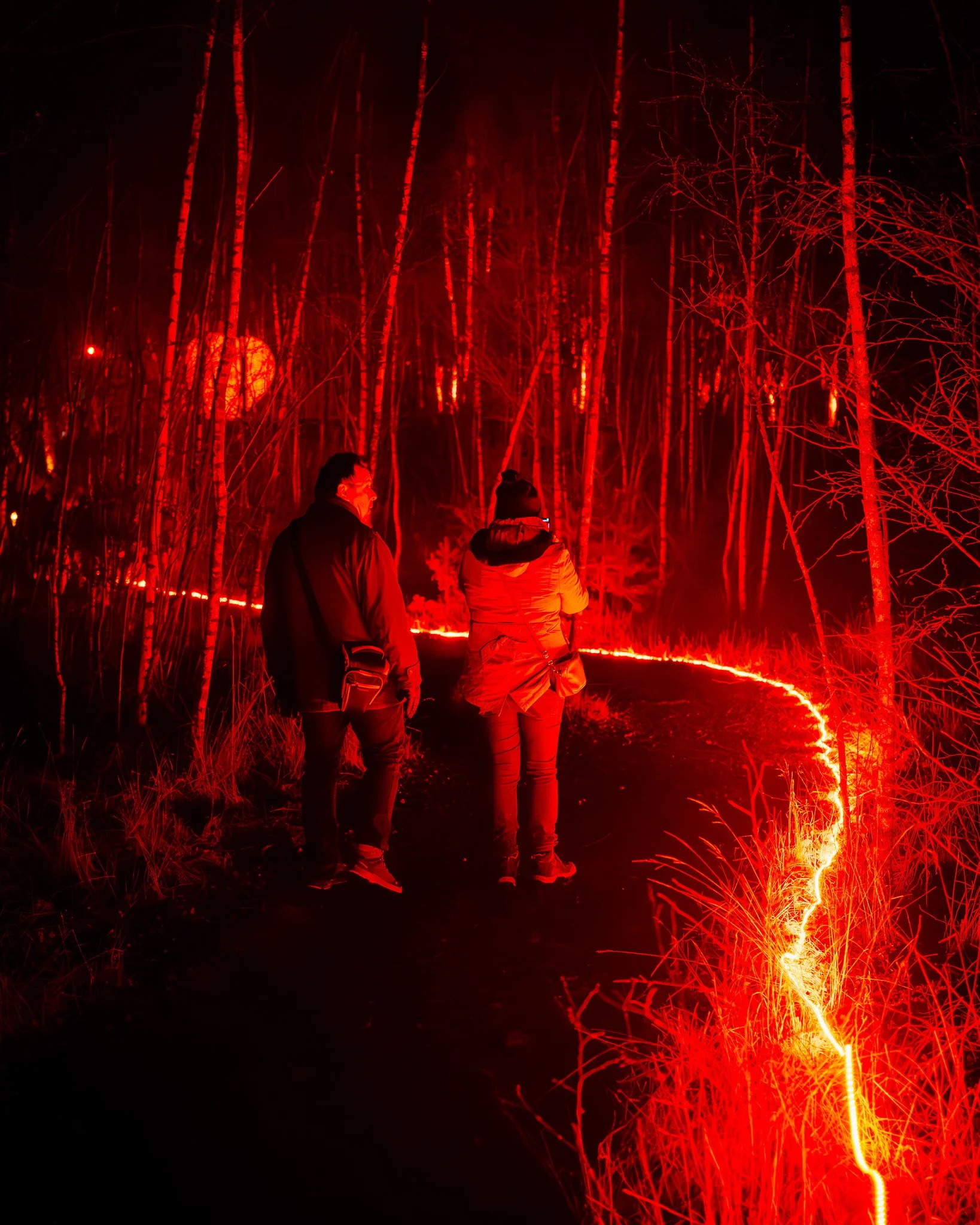 Drie mensen in een bos bij nacht die naar een rood verlichte voorstelling kijken, met vuurwerk op de grond.