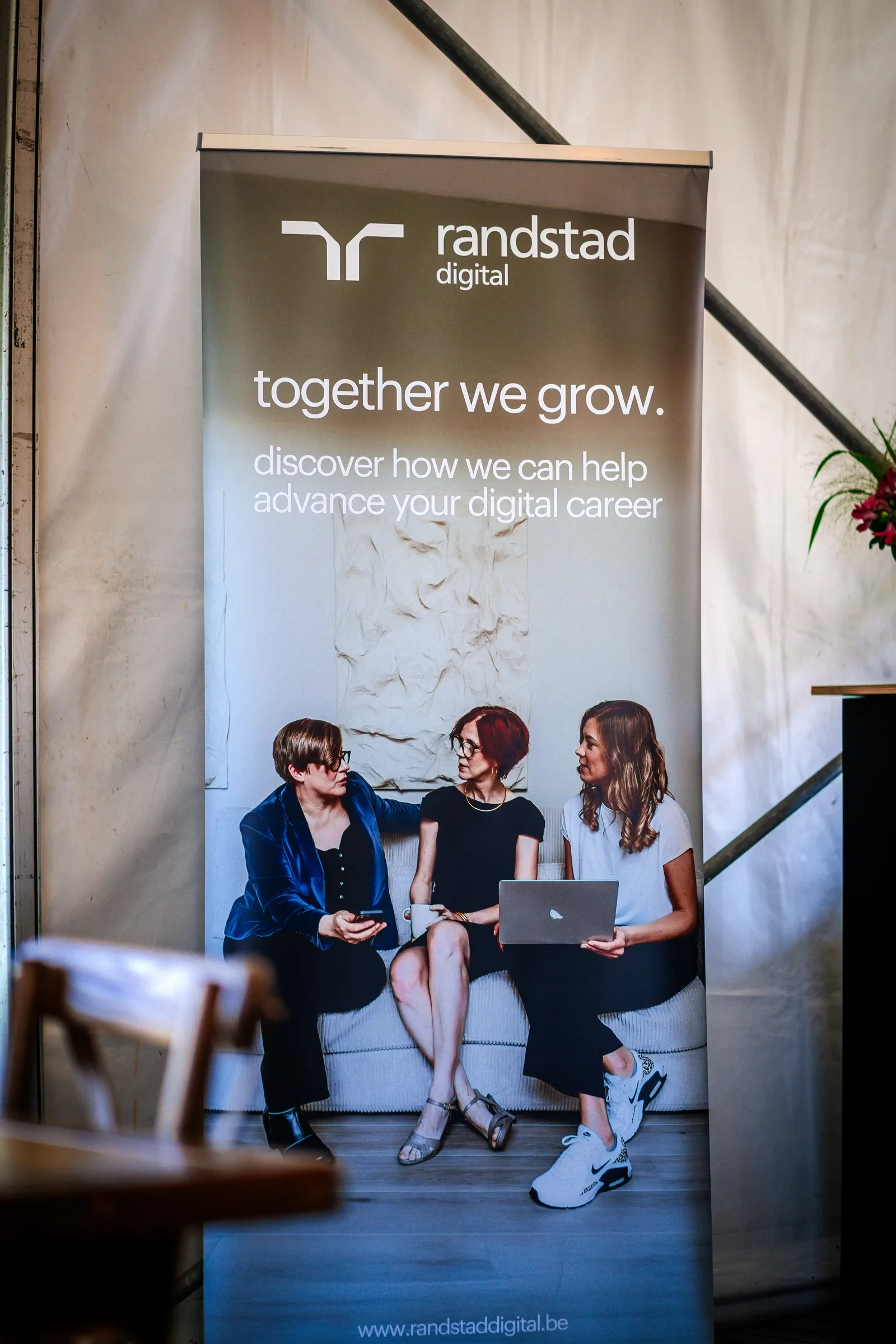 Een promotiebanner van Randstad Digital waarop vier vrouwen in een woonruimte zitten en met elkaar praten, met laptops en een mok in hun handen.