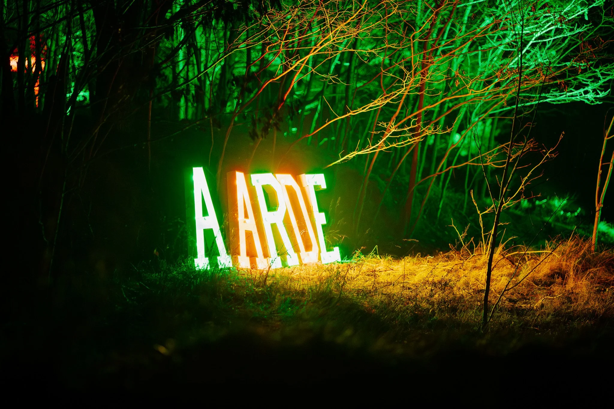 Een verlichte borden met de tekst 'ACRE' staat in een bos, omgeven door groene en oranje verlichting op bomen en struiken.