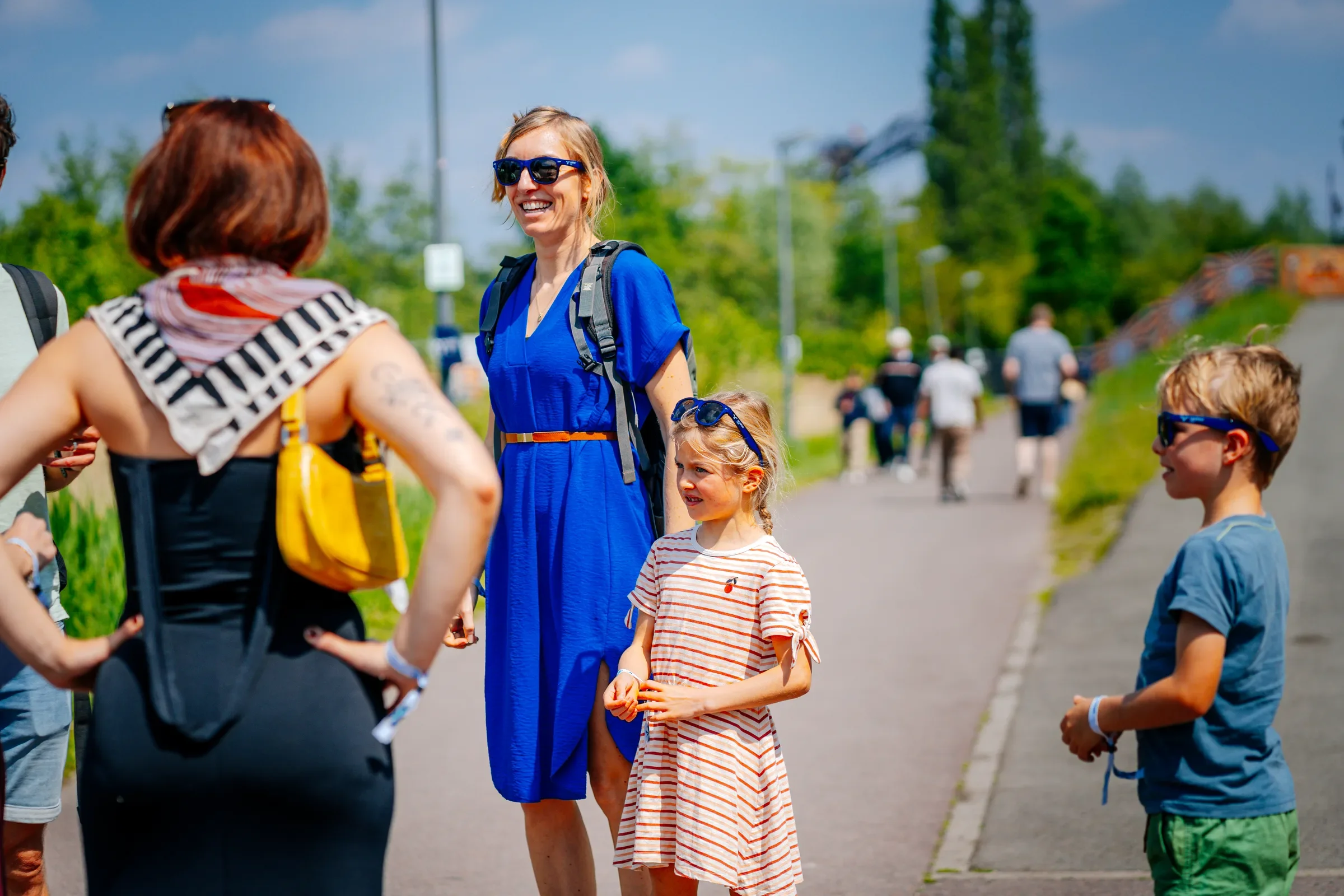 Vrouw in een blauwe jurk en zonnebril die lacht terwijl ze met kinderen en anderen praat op een dag buiten, met groene bomen en een partly cloudy sky op de achtergrond.