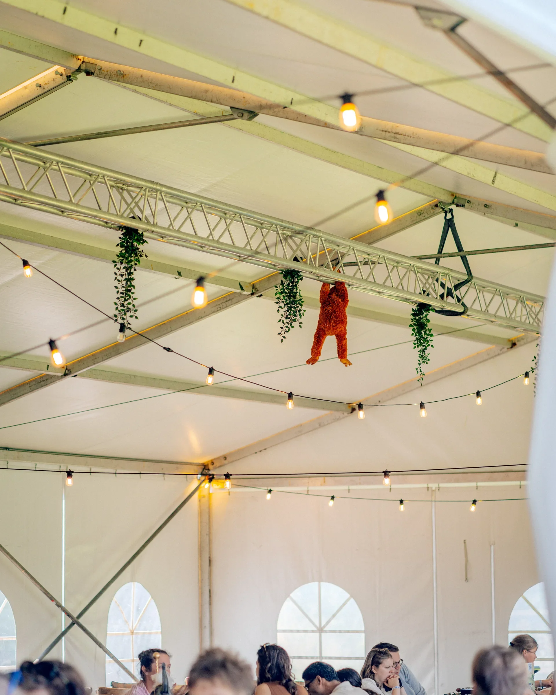 Binnenkant van een grote tent met hangen lichtsnoeren en een metalen constructie aan het plafond. Mensen zitten aan tafels, praten en lezen.