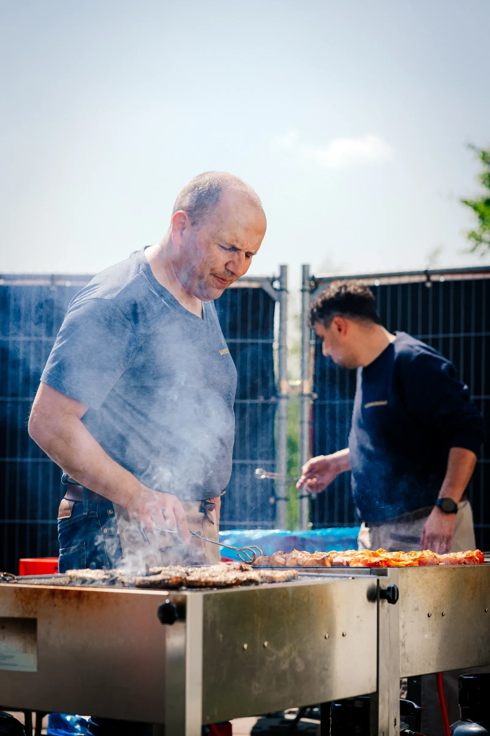 Twee mannen grillen eten op een barbecue buiten, één man kijkt geconcentreerd terwijl hij met tangen werkt, de ander staat op de achtergrond, beiden lijken te genieten.