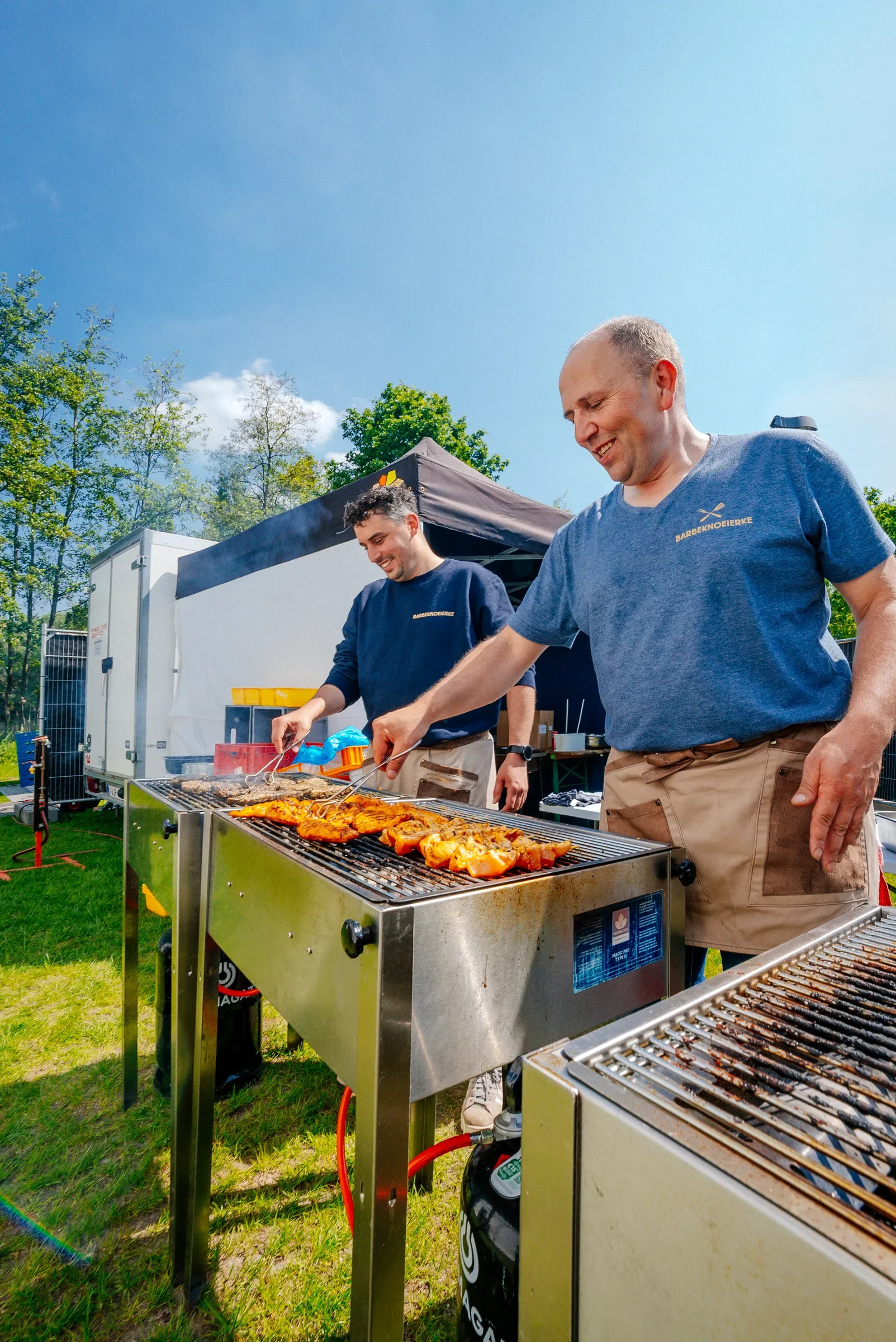 Twee mannen die barbecue gerechten bereiden in een tuin op een zonnige dag. Ze lachen en dragen blauwe shirts met de tekst 'Barbeknoeiferke'.