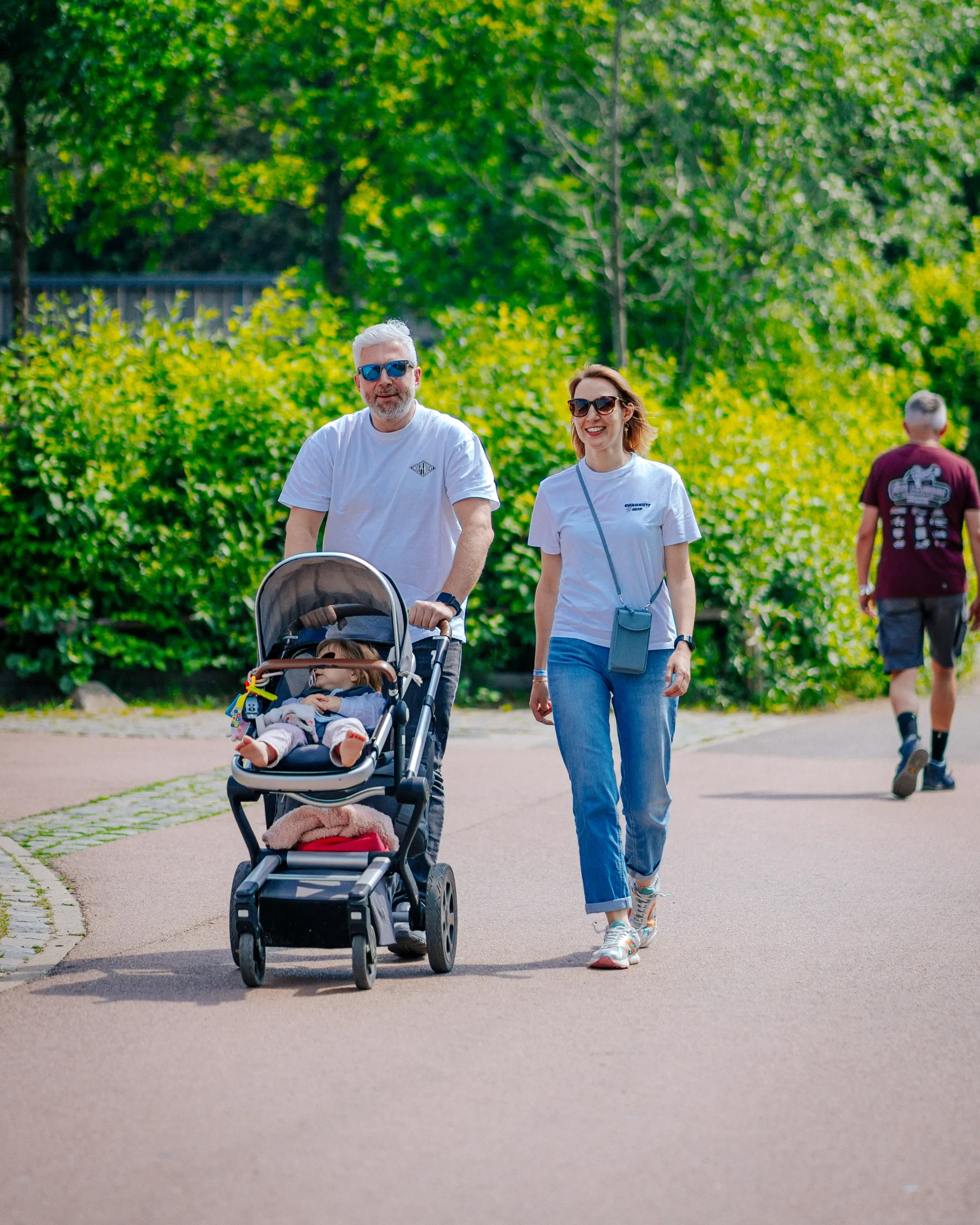 Twee mensen wandelen in een park, een man duwt een kinderwagen met een slapende baby en een vrouw wandelt naast hem lachend. De achtergrond bestaat uit groene bomen en een groep mensen die verder weg lopen.