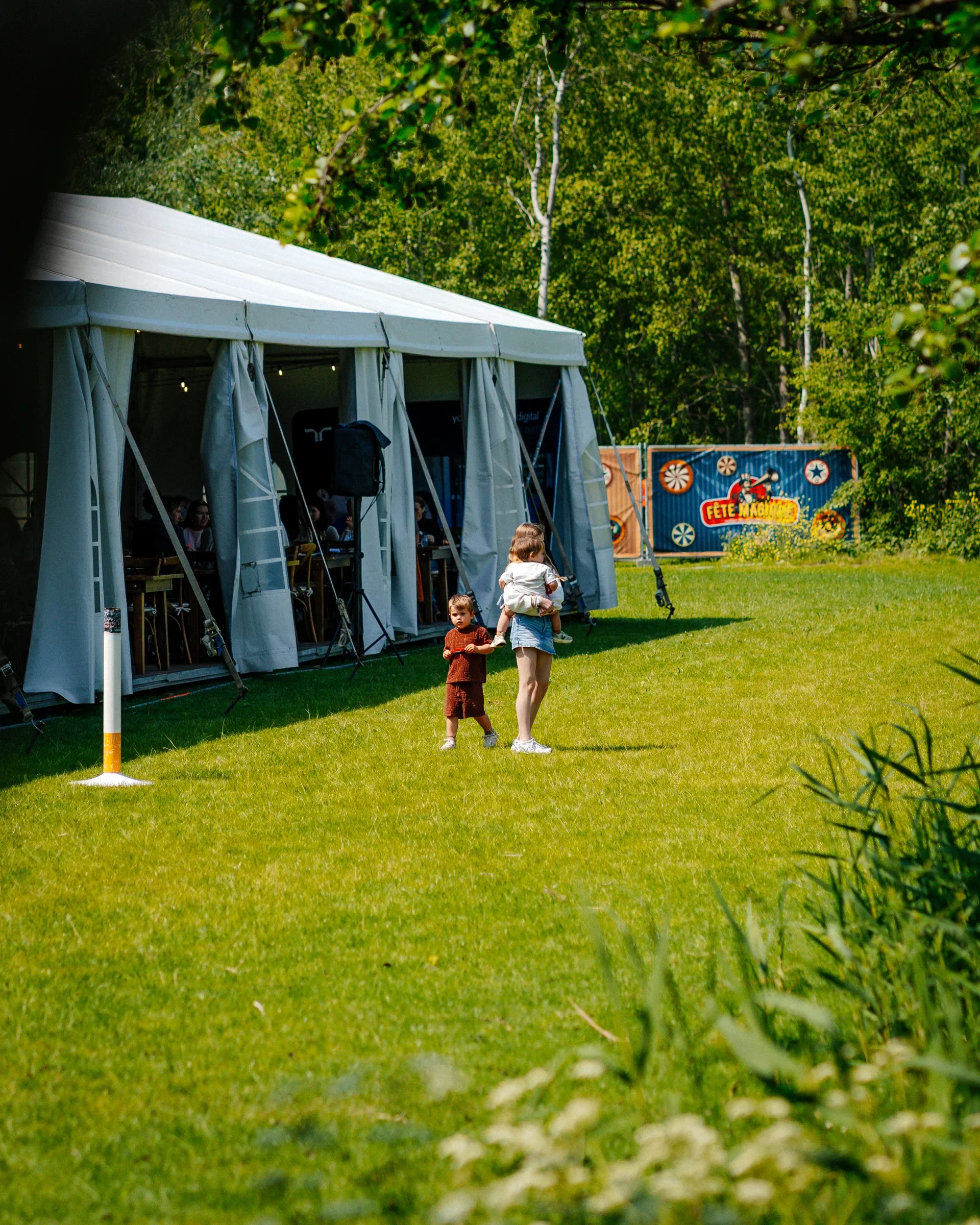 Twee kinderen staan op een grasveld bij een grote witte feesttent met open zijwanden, omringd door groen bos, met een kleurrijke achtergrond met het woord 'Fête Mágique' en decoraties.