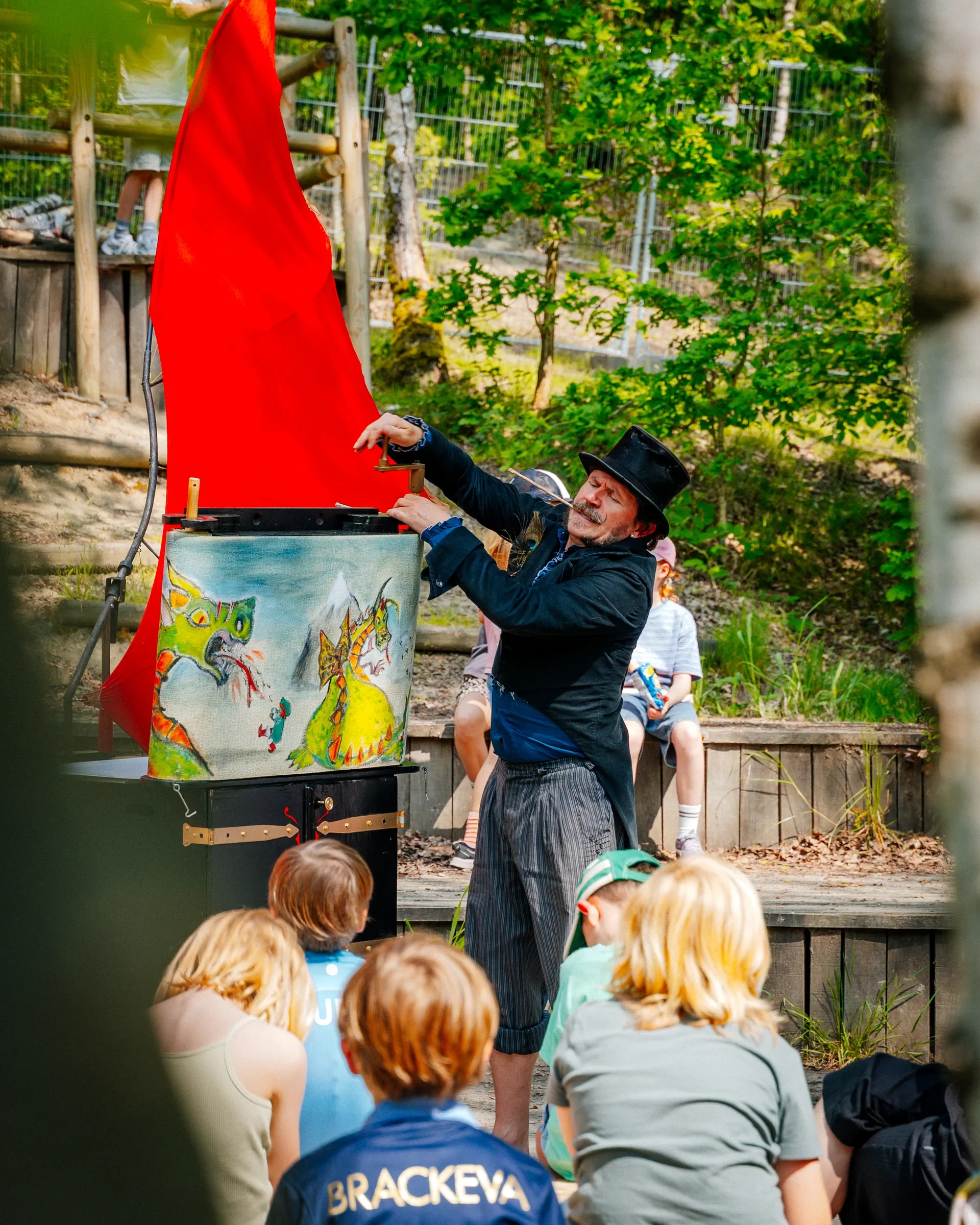 Man verkleedt als tovenaar of circusartiest voert een show uit voor een groep kinderen in een bosrijke omgeving.