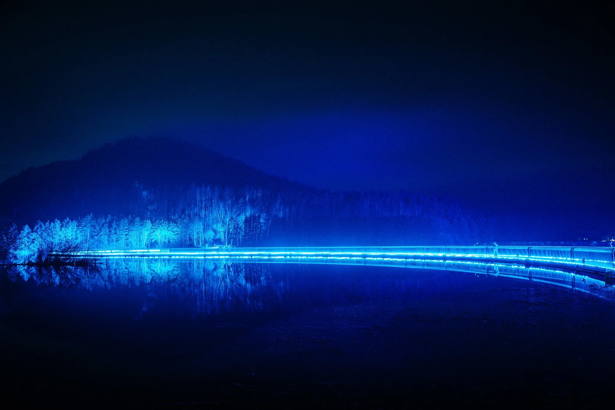 Een nachtlandschap met een rivier, een bos en bergen op de achtergrond, verlicht door blauwe lichten.