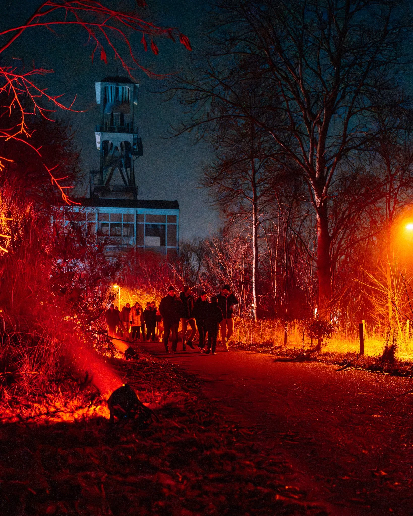 Een groep mensen die een pad bewandelt door een park bij nacht, verlicht door oranje en rode lampen, met kale bomen en een gebouw met een toren op de achtergrond.