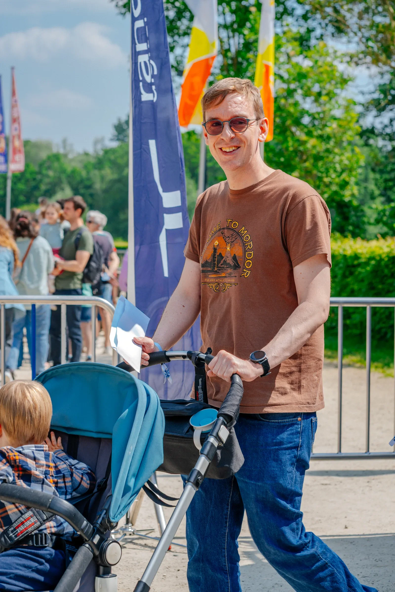 Man met brildraagt en T-shirt met een berglandschap, mensen in de achtergrond op een zonnige dag.