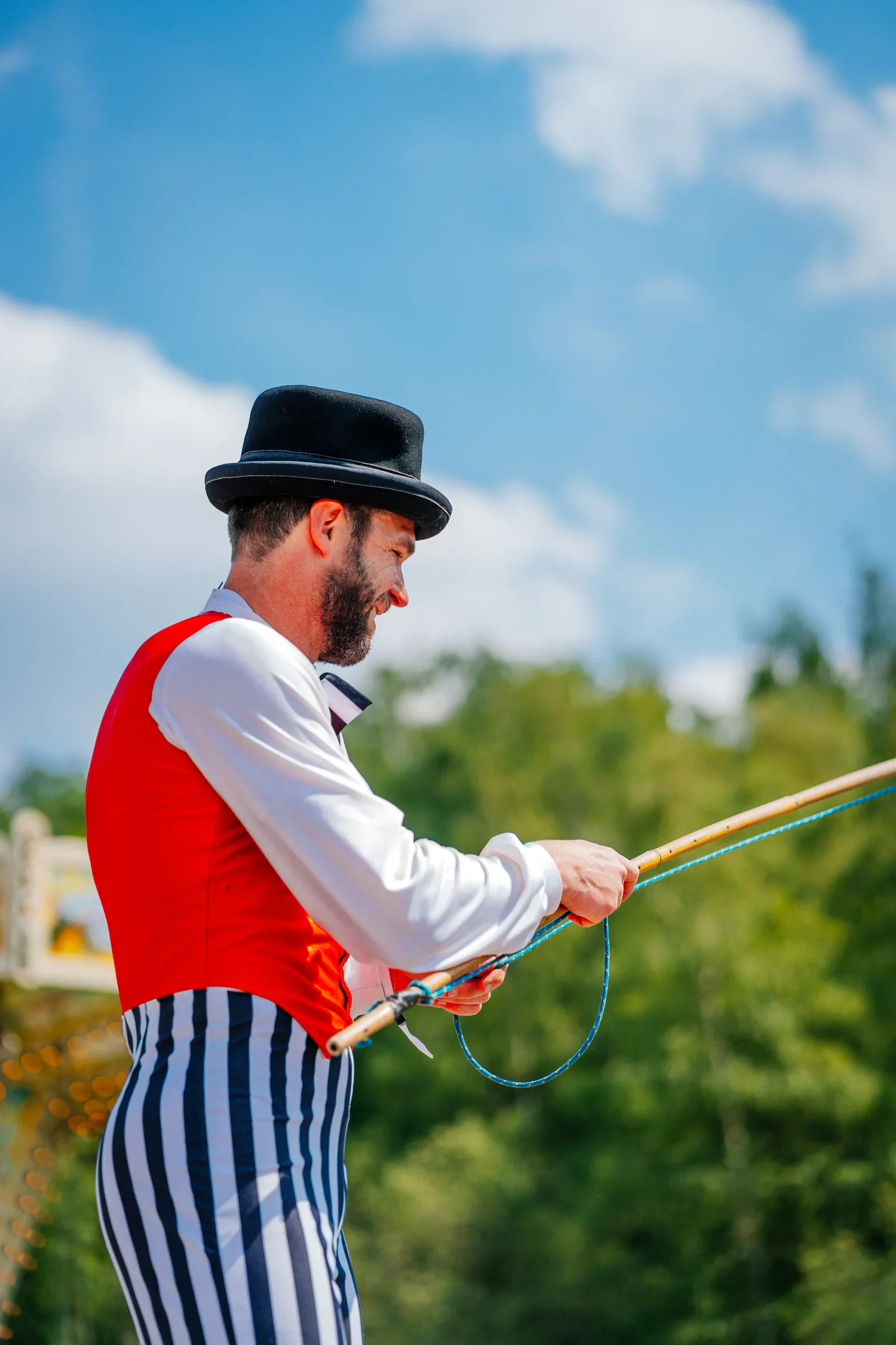 Man in traditionele carnavalskostuum met zwarte hoed en gestreepte broek die een lange stok vasthoudt buiten op een zonnige dag.