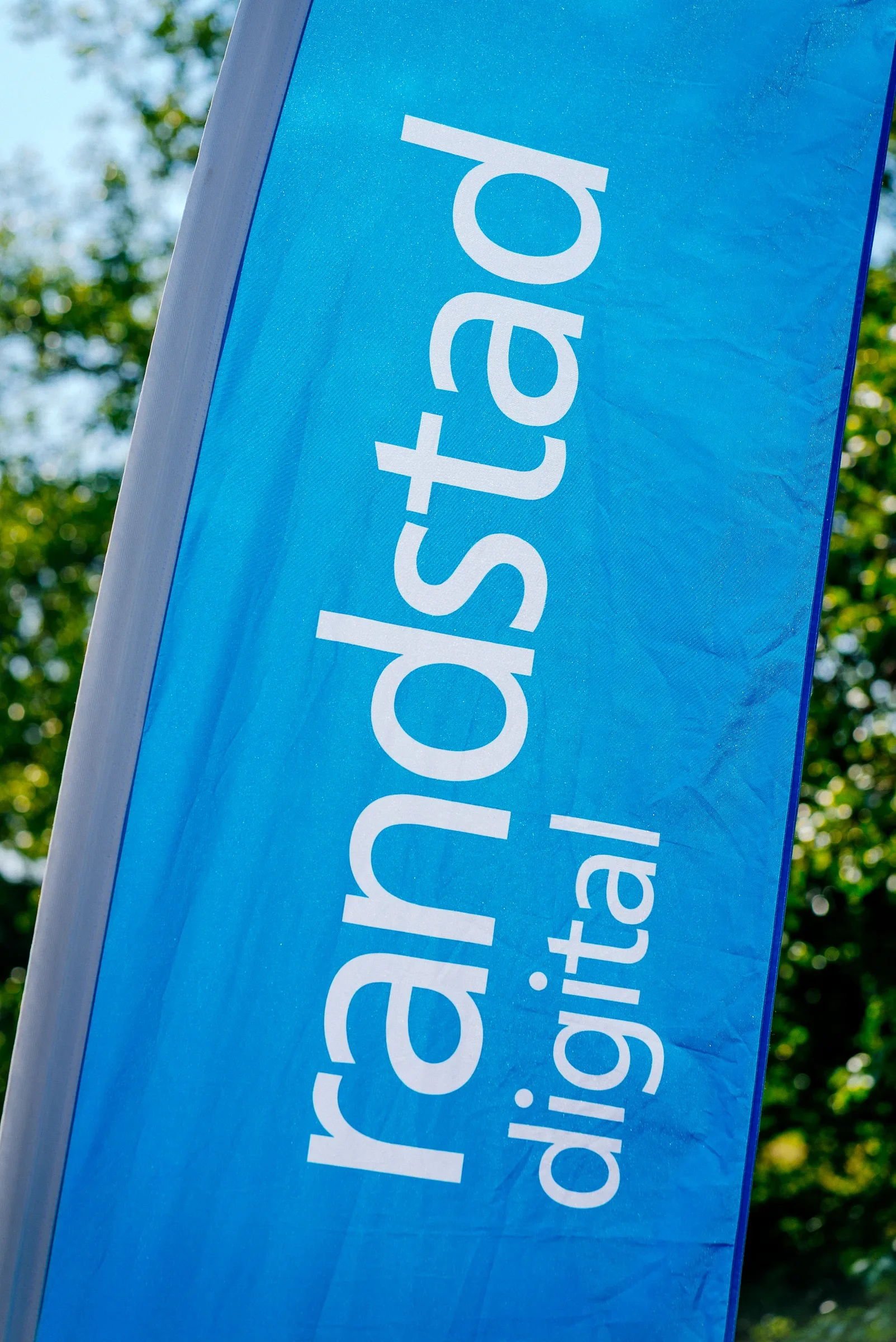 Een blauwe vlag met witte tekst, deels zichtbaar, hangt buiten met een groene bladerige achtergrond en een blauwe lucht.