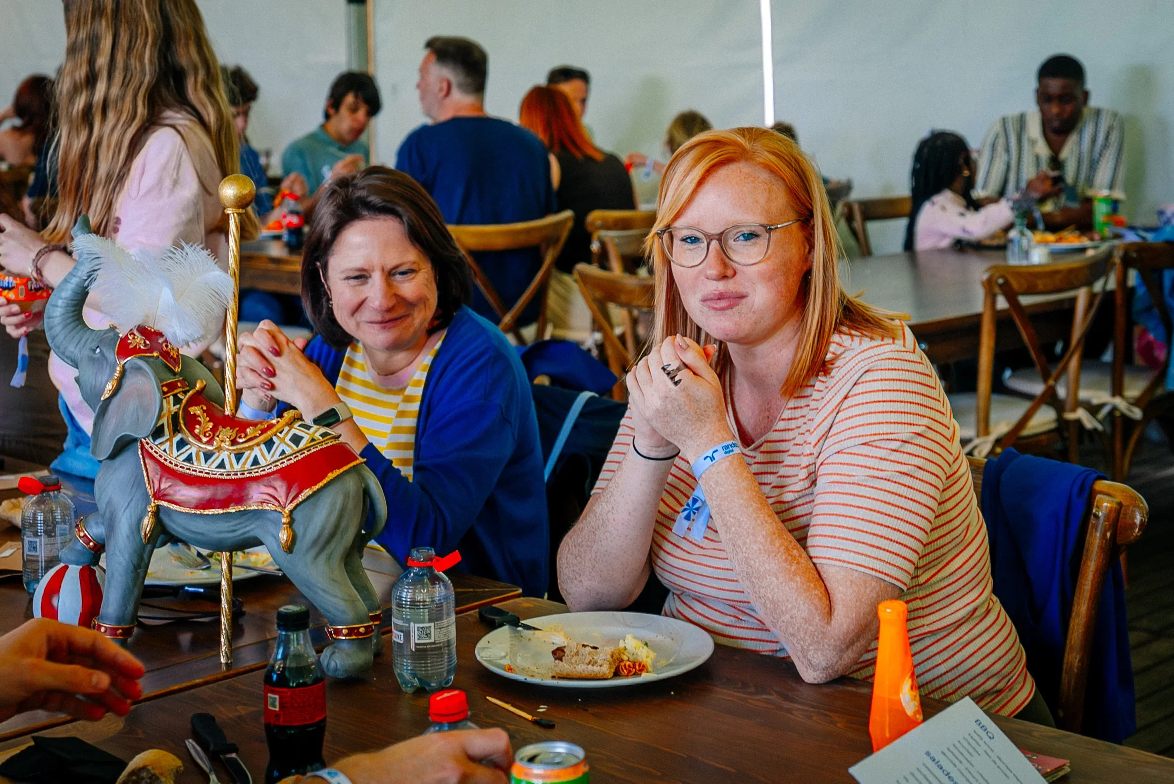 Gezicht van twee vrouwen die aan een tafel zitten, met een decoratief paard en koets tussen hen in, tijdens een sociale bijeenkomst.