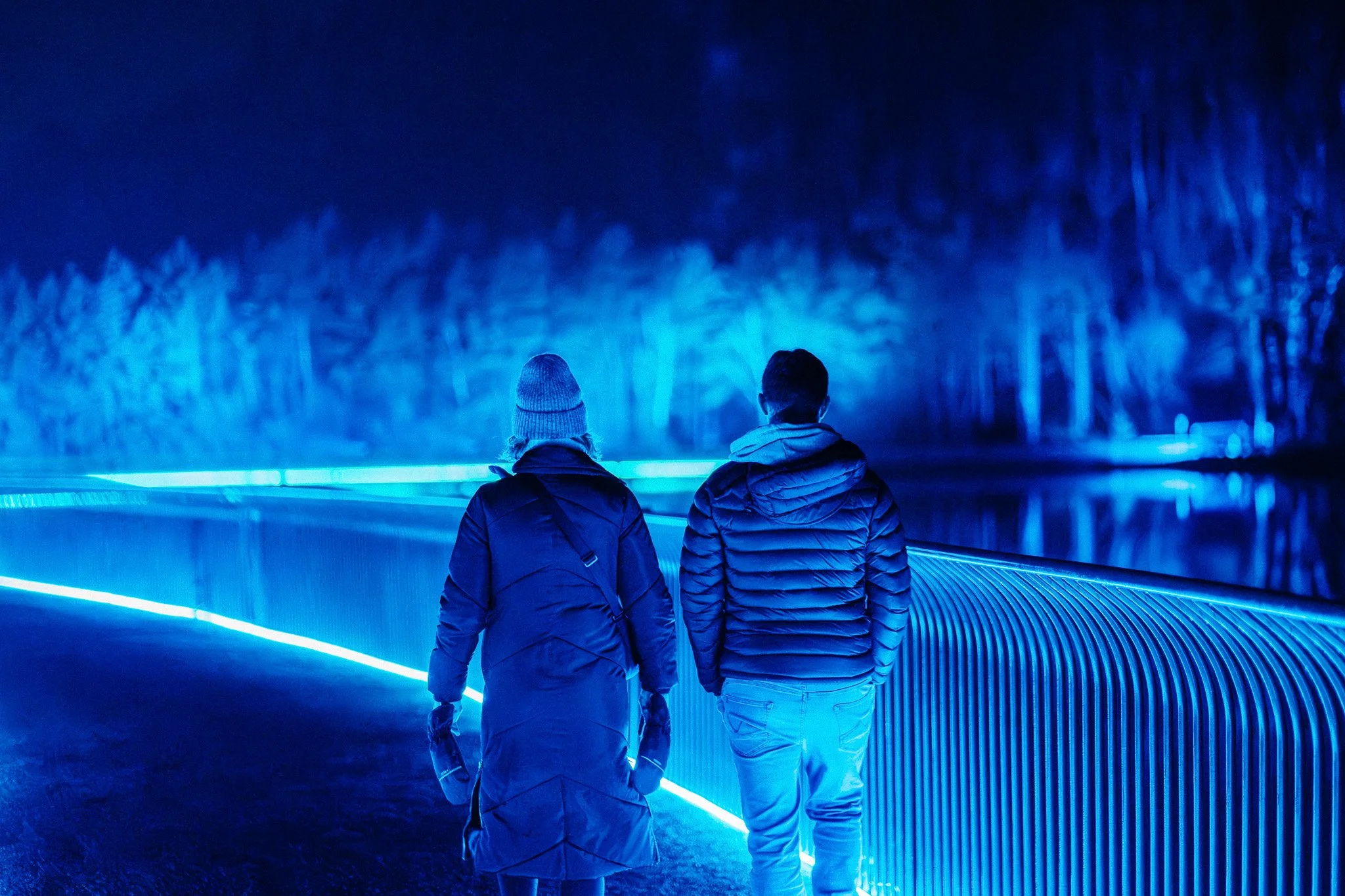 Twee mensen wandelen door een verlichte blauwe tunnel, omgeven door een bos in de achtergrond.