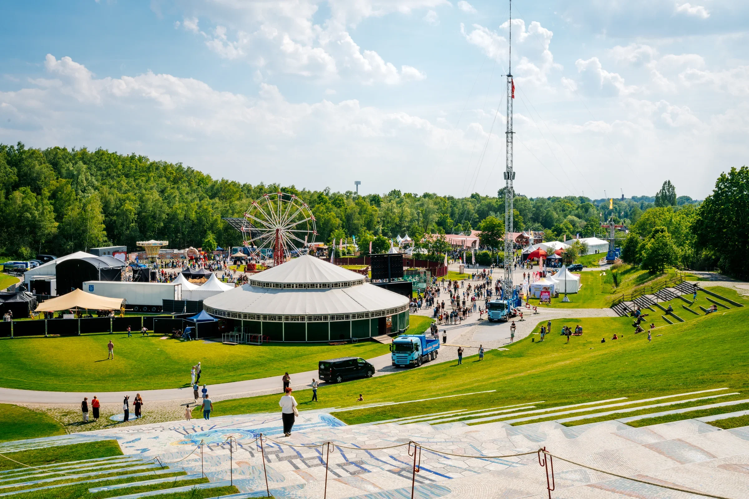 Het foto toont een festivalterrein met een reuzenrad, een grote tent en een mensenmassa onder een heldere blauwe lucht met witte wolken, omringd door groene bomen en een grasveld.