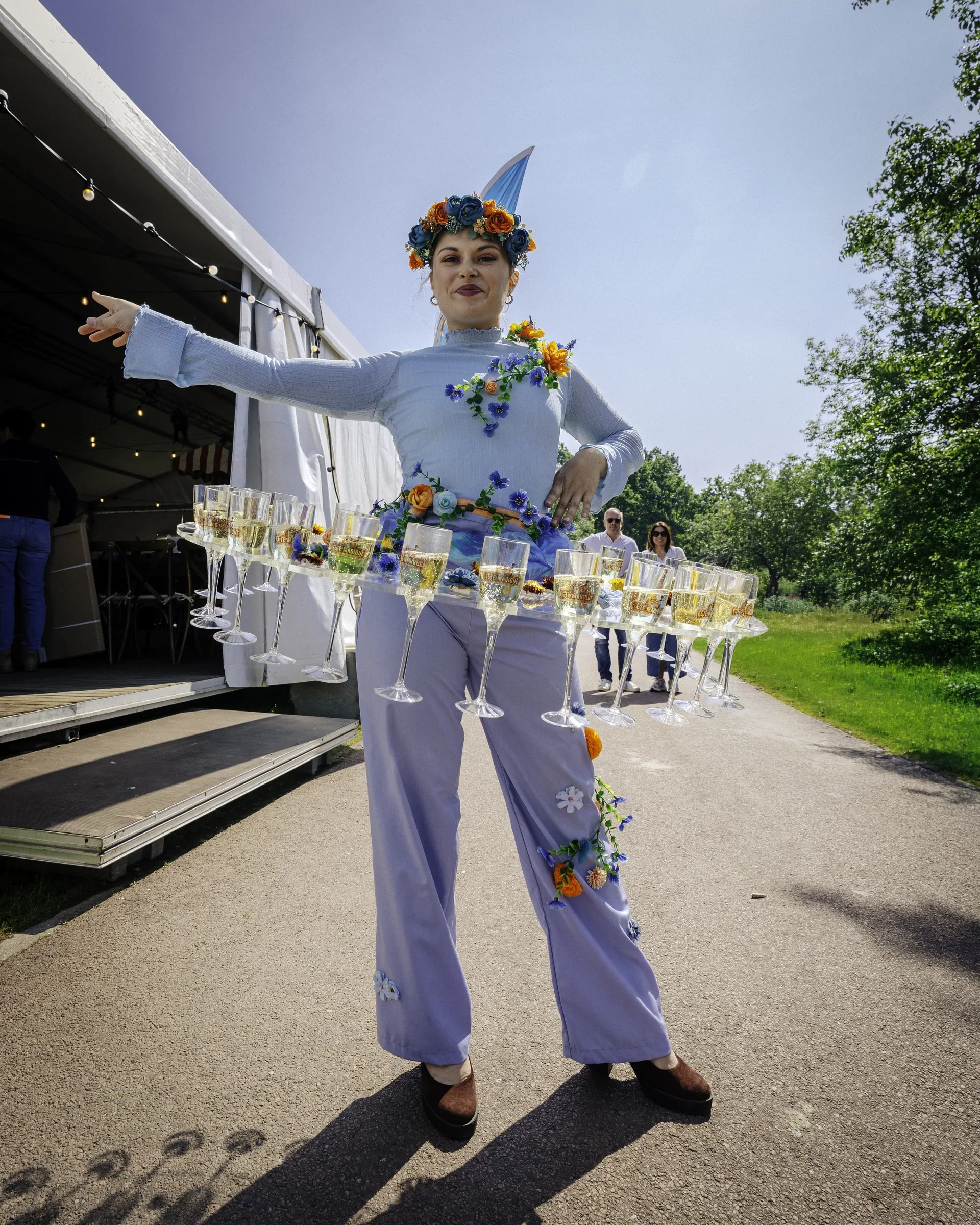 Vrouw in bloemenversiering en een eenhoornhoed die op een dienblad met glazen champagne staat bij een buitenfeest.