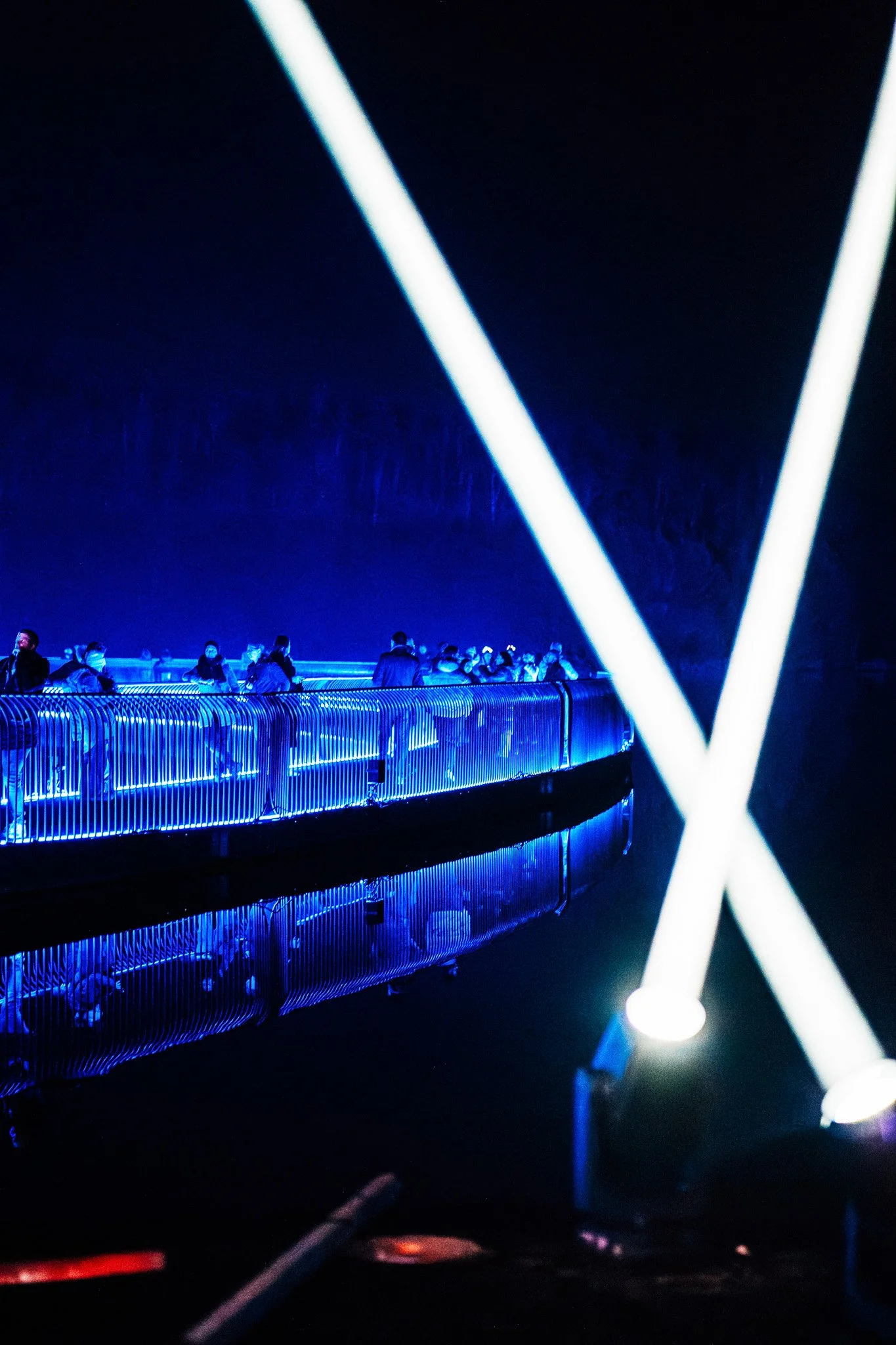 Mensen staan op een brug onder donkere hemel met blauwe en witte neonlichten, op een plek die lijkt op een futuristisch of neon verlicht landschap.