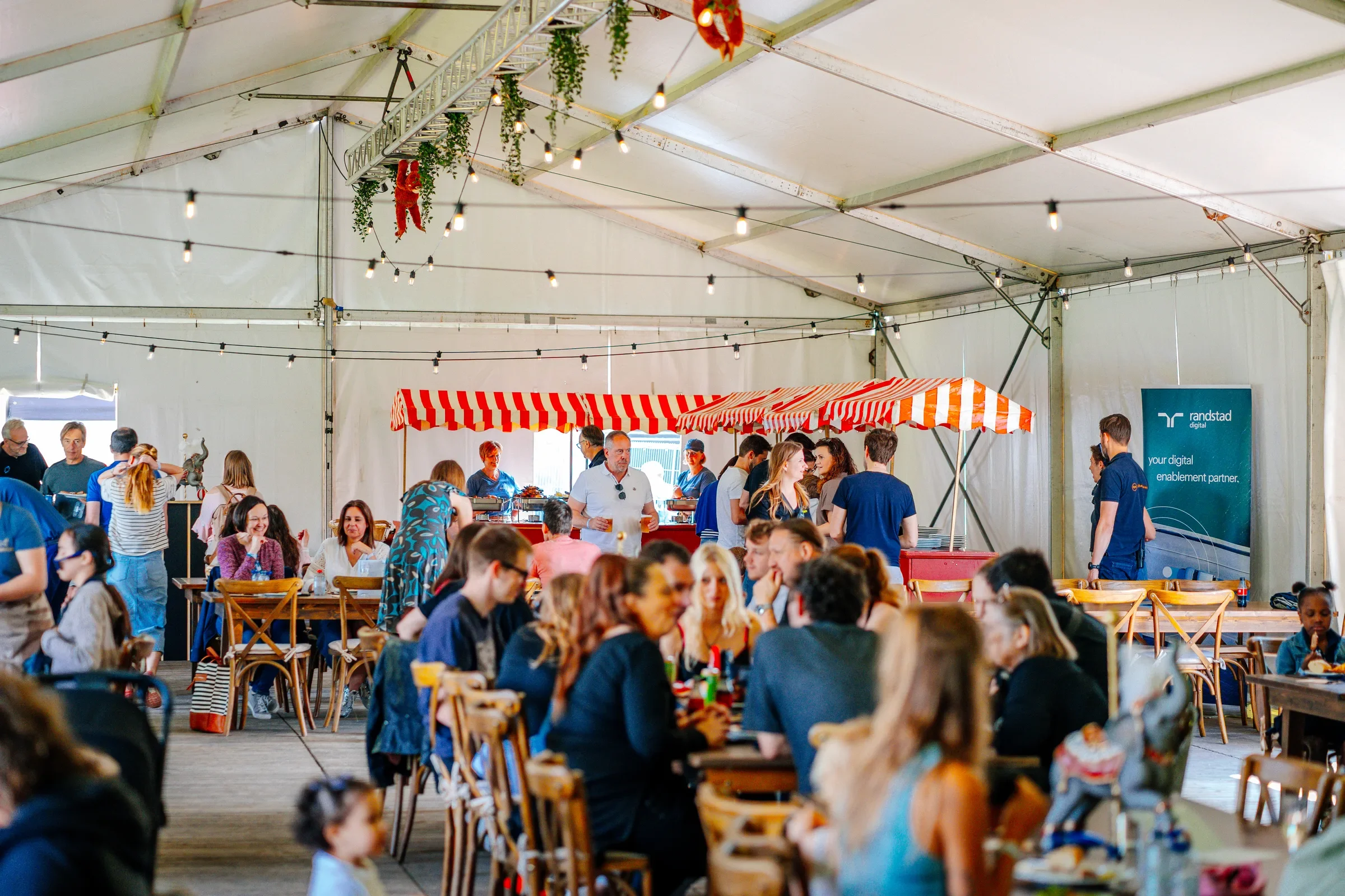 Een drukke binnenmarkt met veel mensen die aan tafels zitten of staan, een tent met rood-witte strepen en decoraties zoals groene planten en hanging lampjes, onder een grote tent met metalen frame en witte zeilen.