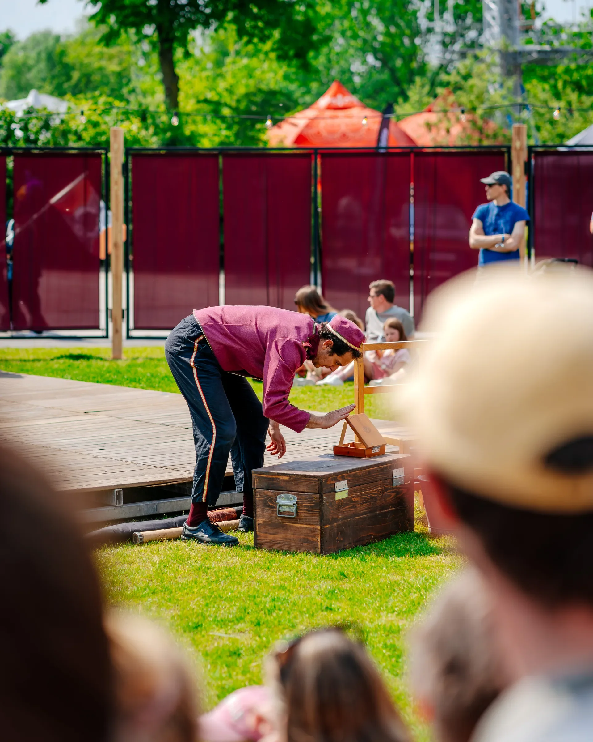 Man buigt voor een houten kist op een podium, omlijst door de hoofden van jonge kinderen, tijdens een buitenoptreden met een publiek van kinderen en een achtergrond van groene bomen en een rode tent op een zonnige dag.