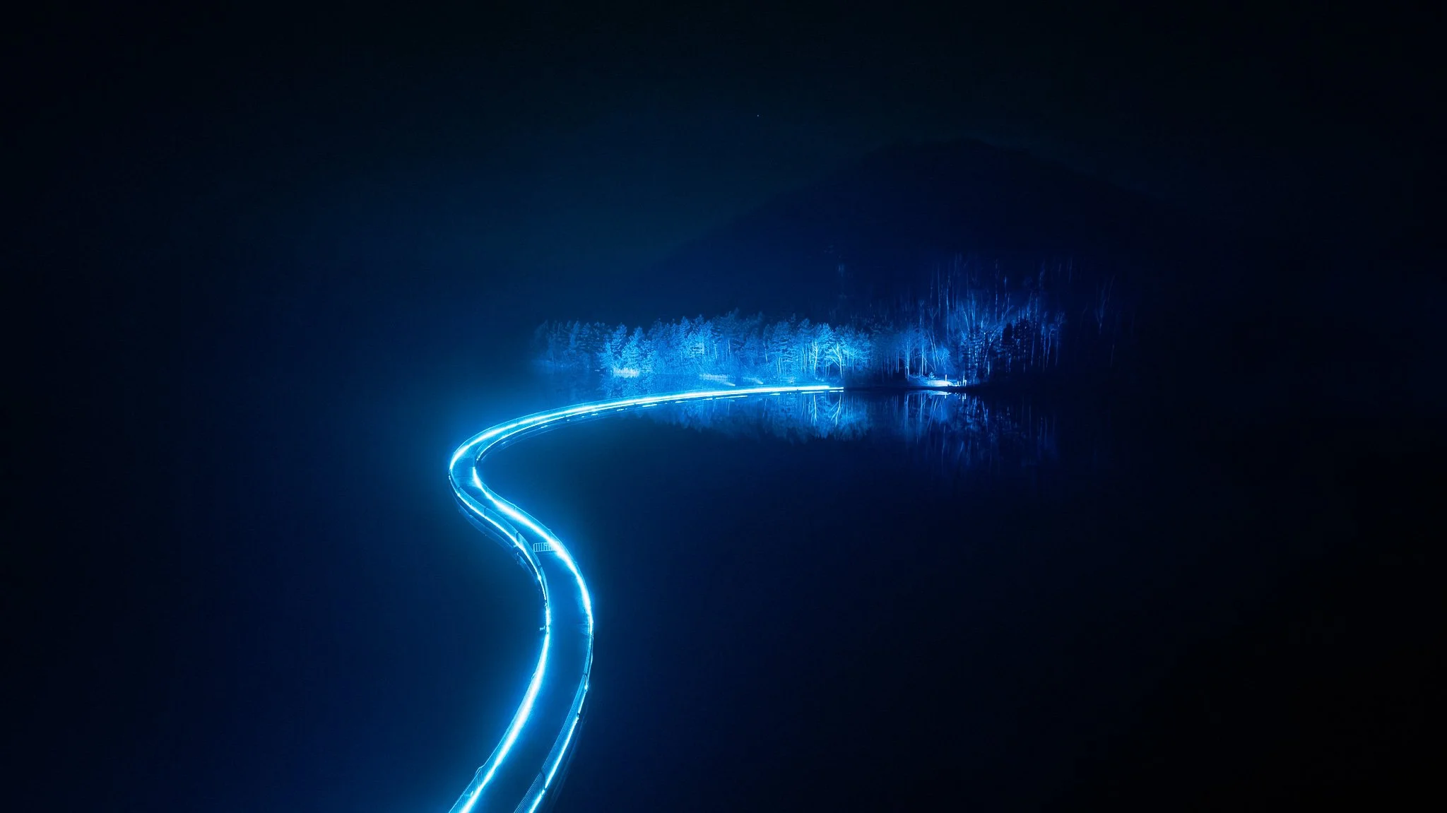Lichtspoor verlicht een weg door een donkere nacht met bomen en een wateroppervlak dat de reflectie van de verlichting toont.